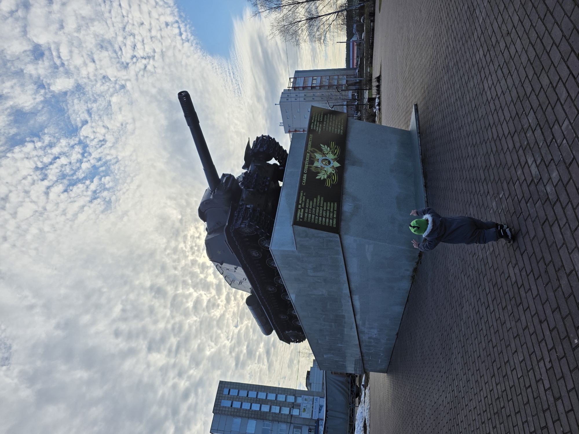 Here is a description of the image:
The image shows a T-34 tank monument in the city of Chaikovsky, Russia. The tank is mounted on a concrete pedestal with a plaque that reads "Glory to Soviet Tankers" and lists the names of soldiers who did not return from the war. A child in a grey snowsuit and green hat stands near the edge of the pedestal. Buildings are visible in the background. The sky is partly cloudy. The ground is covered with brick pavers.