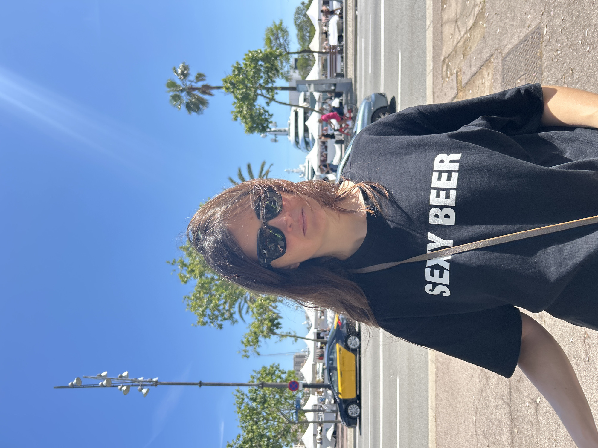A medium shot captures a woman outdoors on a sunny day. She is wearing sunglasses and a black t-shirt with the words "SEXY BEER" printed in bold white letters. A light-colored strap crosses her torso. In the background, the scene unfolds along a city street with palm trees, street lights, and trees. There is a yellow and black taxi, a boat, and white tents or structures. The location appears to be in Barcelona, Spain.