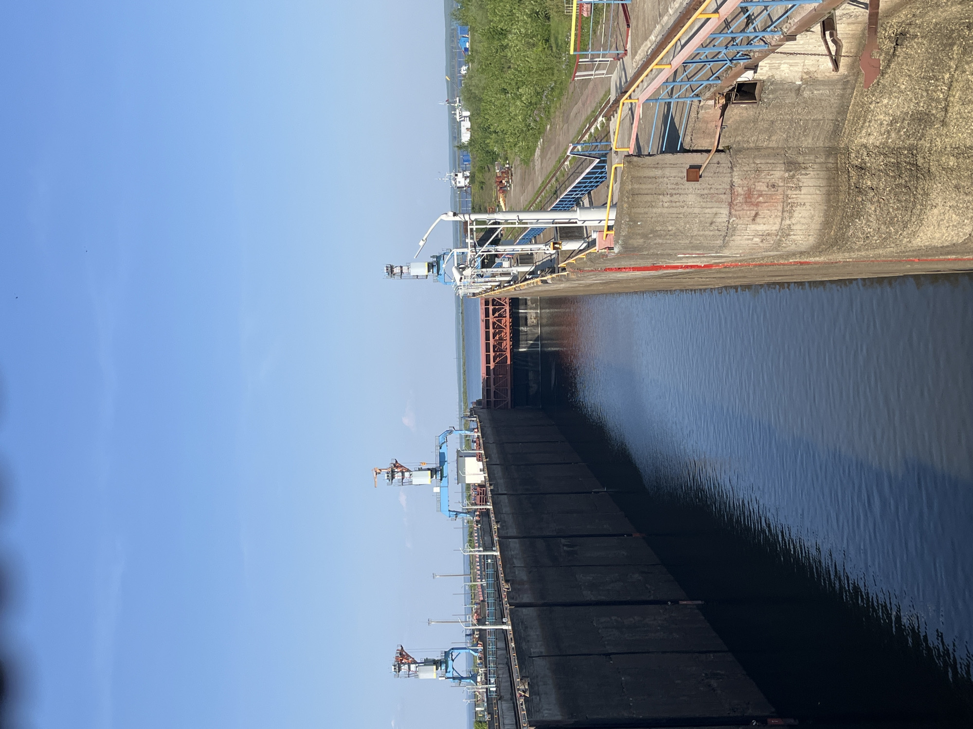 The image depicts a canal lock on the Kama River in Chaikovsky, Russia. The lock walls are made of concrete, with metal walkways and railings along the top. The lock gate is black and has metal structures on top. The water in the lock is dark and reflects the sky. In the background, there are trees and buildings. There are also white ships docked in the distance. The sky is clear and blue.