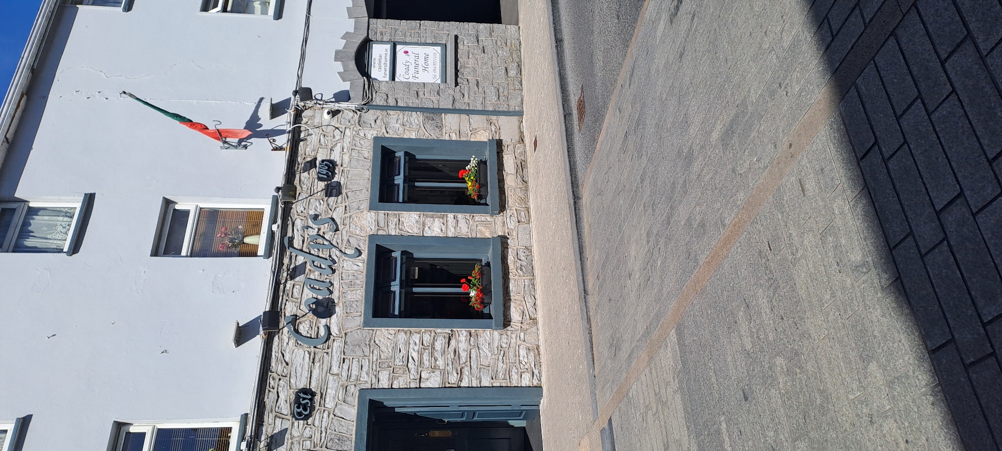 The image shows the exterior of a building with a stone facade. The building has several windows, some with flower boxes. A sign on the building reads "Coady's Funeral Home" and includes a website address "www.castlebarfuneralhome.ie". Another sign appears to say "Est 1905". The building has a white upper section and a grey stone lower section. A flag, possibly the Irish flag with an added green element, is mounted on the wall. The scene appears to be outdoors on a sunny day, with shadows cast on the pavement in front of the building. The pavement is made of stone slabs. The overall impression is of a historic building in a town.