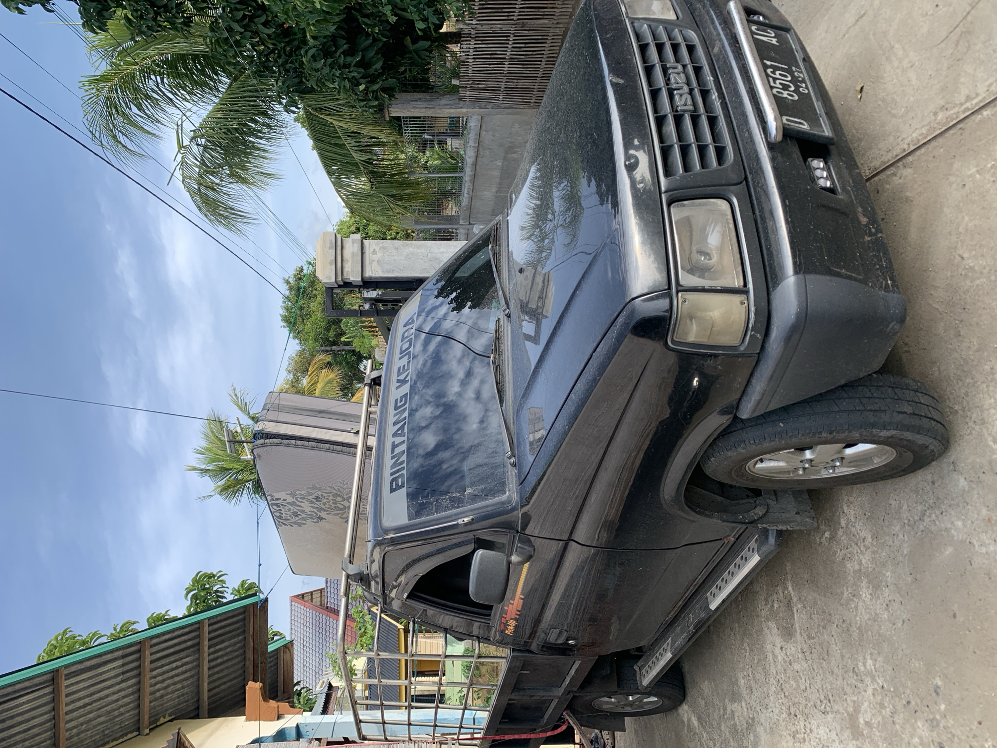 A black Isuzu pickup truck is parked on a concrete surface in front of a residential building in Meunasahdrang, Indonesia. The truck has a metal cage in the bed and a mattress strapped to the roof rack. The license plate reads "D 8561 AC". The building to the left has a corrugated metal roof and a light-colored facade. Tropical trees and a fence are visible in the background, under a cloudy blue sky.
