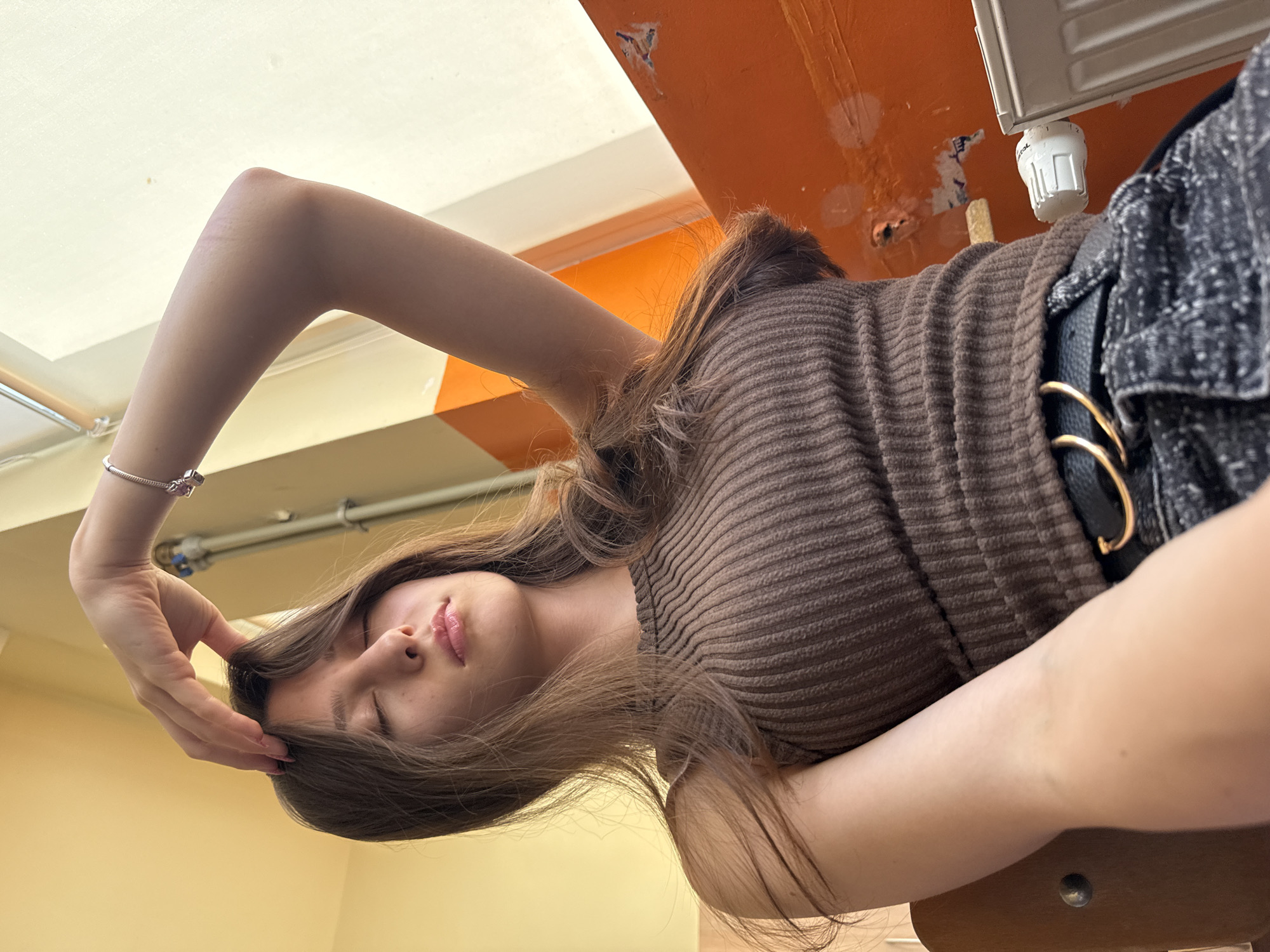 A young woman with long brown hair is sitting indoors, possibly in a classroom or an institutional setting. She has her eyes closed and her right hand is raised, touching her forehead or hair. She is wearing a brown ribbed sleeveless top and dark jeans with a belt featuring two gold rings. The background shows a wall painted in yellow and orange, with a white radiator visible on the right. A pipe and what appears to be a window frame are also present. The lighting suggests it's daytime, possibly near a window. The location is identified as Sofia, Bulgaria.