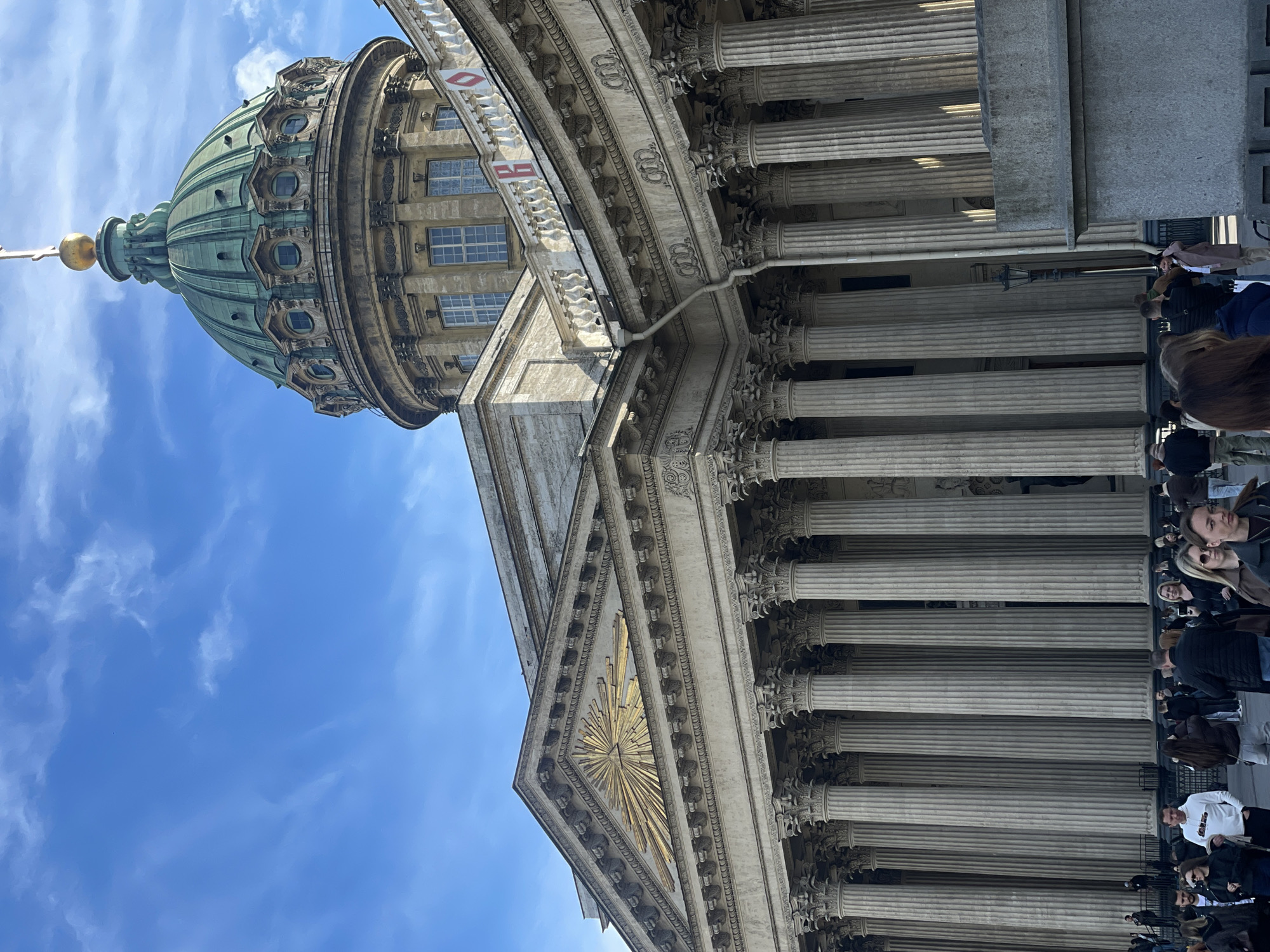 The image presents a striking, upward view of the Kazan Cathedral in Saint Petersburg, Russia, bathed in bright daylight under a clear blue sky dotted with scattered white clouds. The grand Neoclassical architecture of the cathedral dominates the scene, highlighting its imposing scale and intricate details.
The prominent, green-patinated copper dome is a central feature, exhibiting a ribbed structure, circular windows, and ornate embellishments. A golden finial, likely a sphere and cross, crowns the dome, glinting in the sunlight. Below, a vast colonnade forms the main facade, composed of numerous tall, fluted columns topped with richly detailed Corinthian capitals. These columns create a rhythmic and grand entrance.
Within the triangular pediment above the entrance, a dazzling golden sunburst emblem, possibly representing the Eye of Providence, radiates outwards, adding to the cathedral's symbolic and aesthetic richness. The building's surfaces are adorned with intricate carvings and decorative friezes, showcasing fine craftsmanship.
At the base of the columns, numerous individuals are visible, appearing to be visitors or tourists. They are dressed in casual wear, some looking up at the towering structure, others moving along, suggesting a lively public space. The scene conveys a sense of awe and admiration for the historical and architectural magnificence of the cathedral. A small, indistinct white sign with red text, possibly "MB" or a similar abbreviation, is barely discernible on a balustrade in one of the building's higher sections. The clear, bright weather suggests a pleasant day for sightseeing.