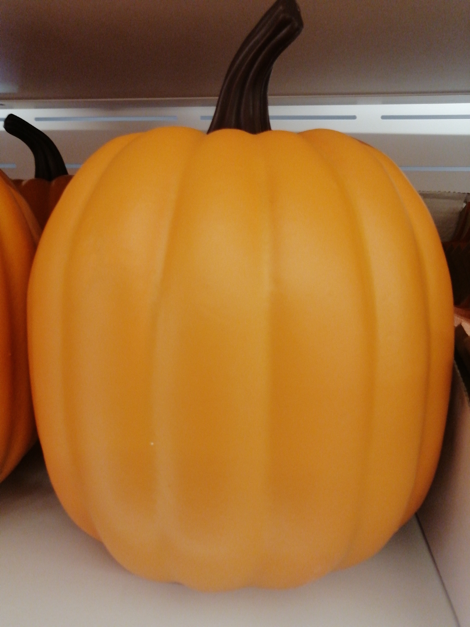 Two artificial pumpkins are displayed on a shelf. The larger pumpkin in the foreground is orange with vertical indentations and a dark brown stem. To the left of the larger pumpkin, a portion of another orange pumpkin is visible, also with a dark stem. The background consists of a light-colored shelf with horizontal white lines. The image is a medium shot, taken from a slightly low angle, focusing on the pumpkins. The setting is indoors, likely a retail environment.