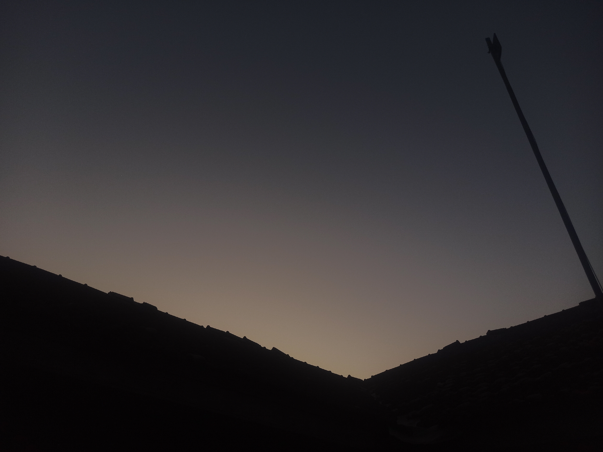 The image depicts a silhouetted view of two tiled rooftops against a dusky sky. The sky transitions from a dark grey at the top to a lighter, yellowish-grey near the horizon. A tall, thin pole with a fork-like structure at its apex is positioned on the right side of the frame, angled upwards against the sky. The rooftops are dark and indistinct, with the texture of tiles visible along their edges. The scene appears to be an outdoor environment in the city of Tuba, Indonesia, likely captured during twilight. There are no people or animals visible in the image.