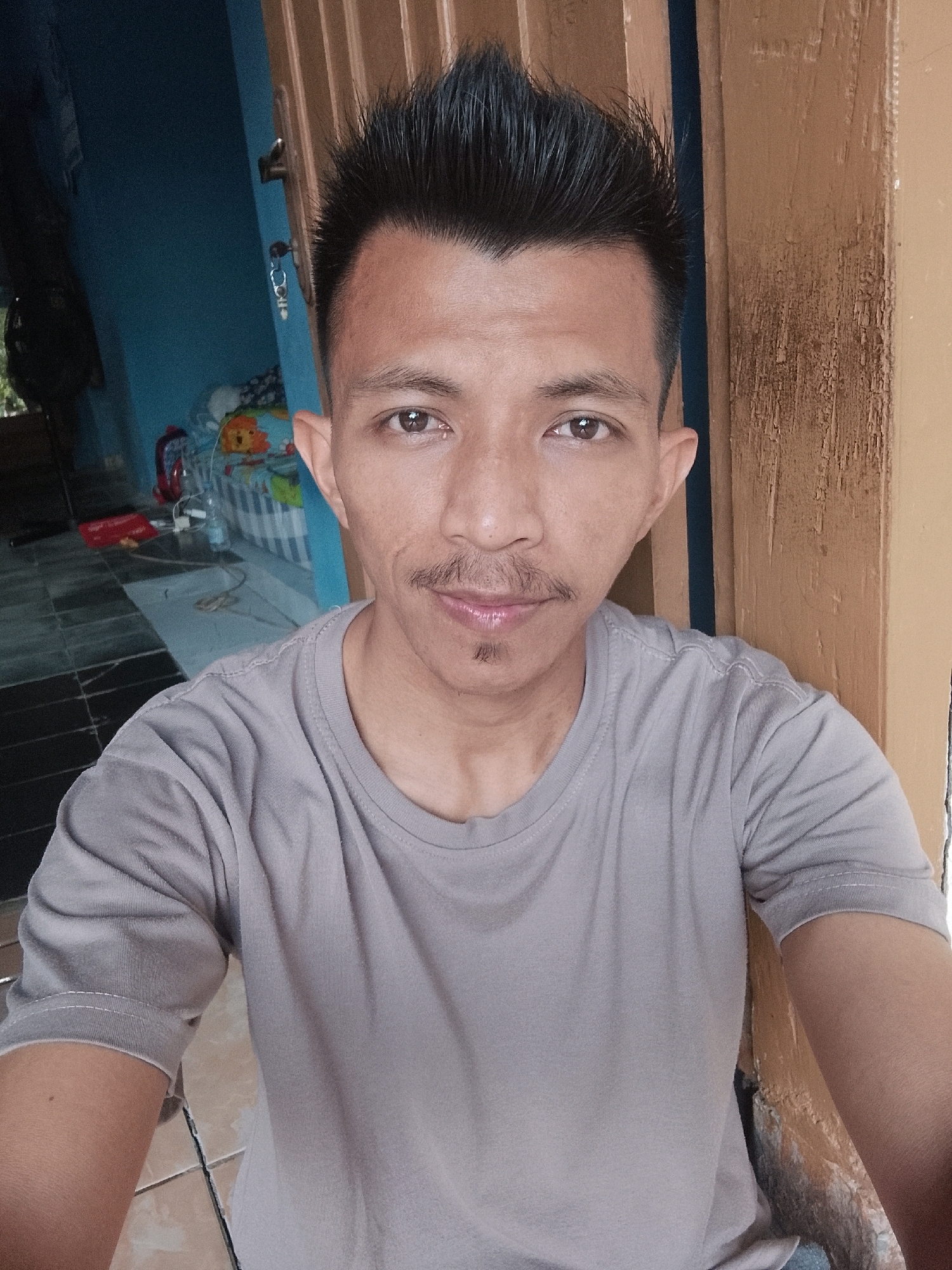 A young man with spiky black hair and a light mustache is pictured in a selfie taken outdoors. He is wearing a plain gray t-shirt. The background includes a partially open wooden door, a blue wall, and a tiled floor. Some objects are visible in the background, including a red object on the floor and a striped basket with some items inside. The scene appears to be in Kertahayu, Indonesia.