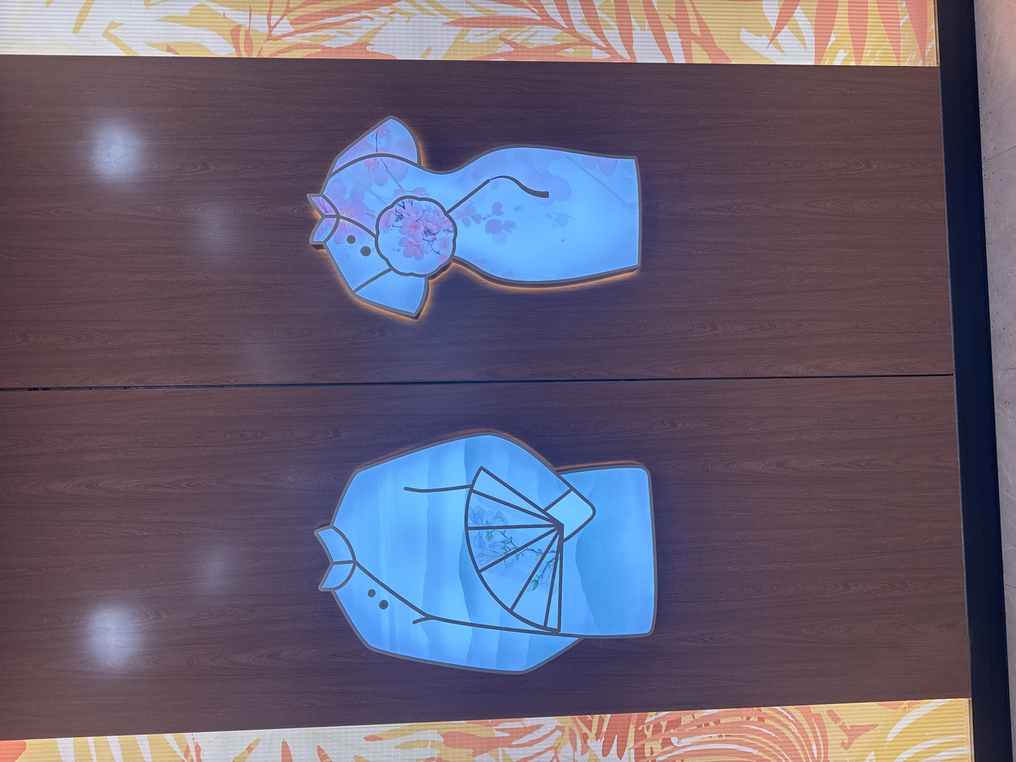 The image shows two signs on a wood-paneled surface. The top sign features a stylized image of a woman's kimono with a cherry blossom pattern. The bottom sign depicts a stylized image of a man's kimono holding a fan. These signs are likely designating the entrances to women's and men's restrooms, respectively. The location is Baiyun District, China.