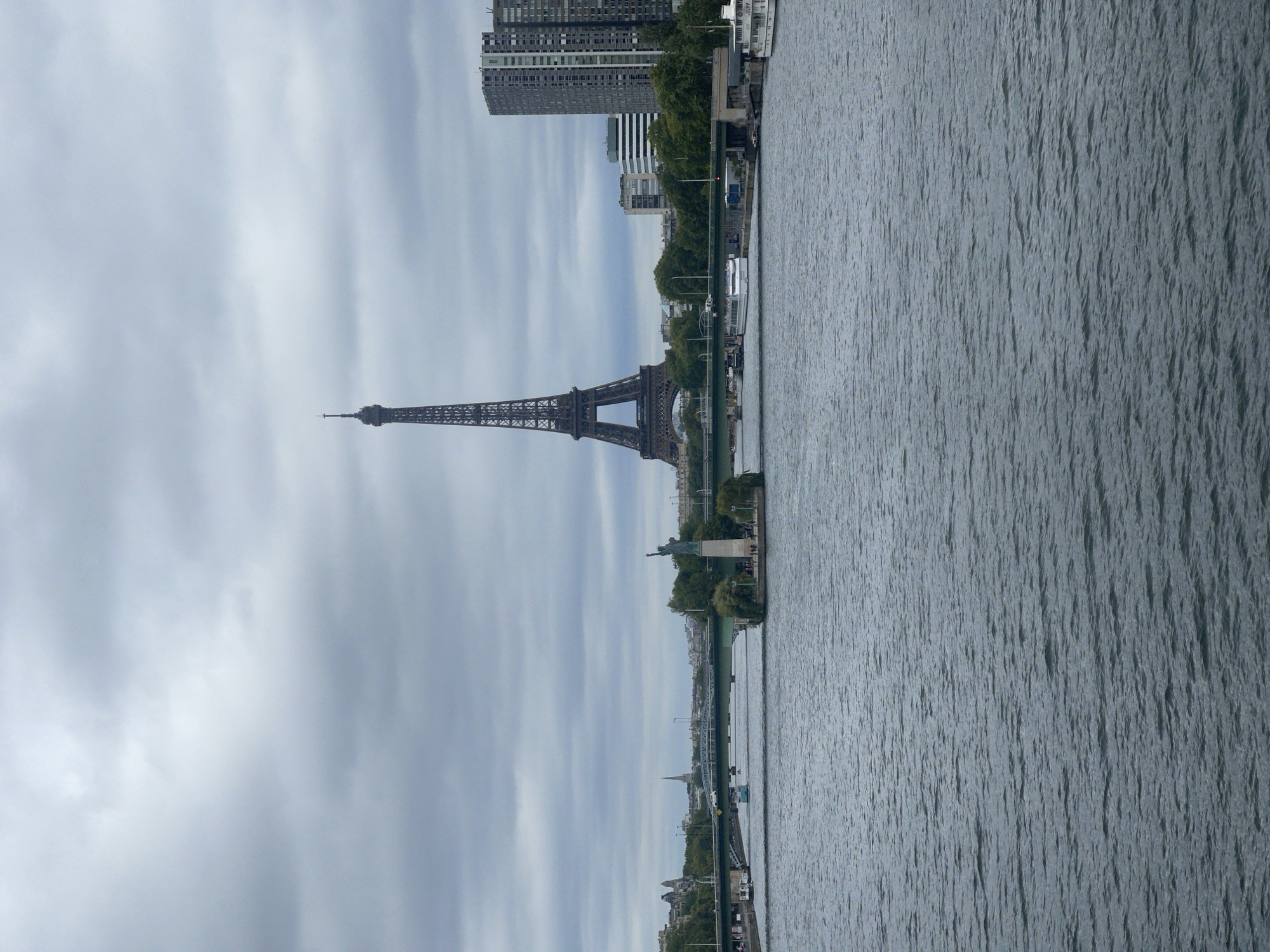 This image captures a view of the Seine River in Paris, France, under an overcast sky. Dominating the background is the iconic Eiffel Tower, its lattice structure clearly visible. In the middle ground, a small island in the river holds a miniature replica of the Statue of Liberty. Several bridges span the river, with traffic and boats visible on them and the water. On the right bank, modern high-rise buildings stand tall, contrasting with the historical architecture on the left bank. The river itself is choppy, reflecting the cloudy sky. There are no discernible people engaged in specific actions, nor are there any direct interactions depicted, creating a serene, albeit slightly somber, urban landscape.