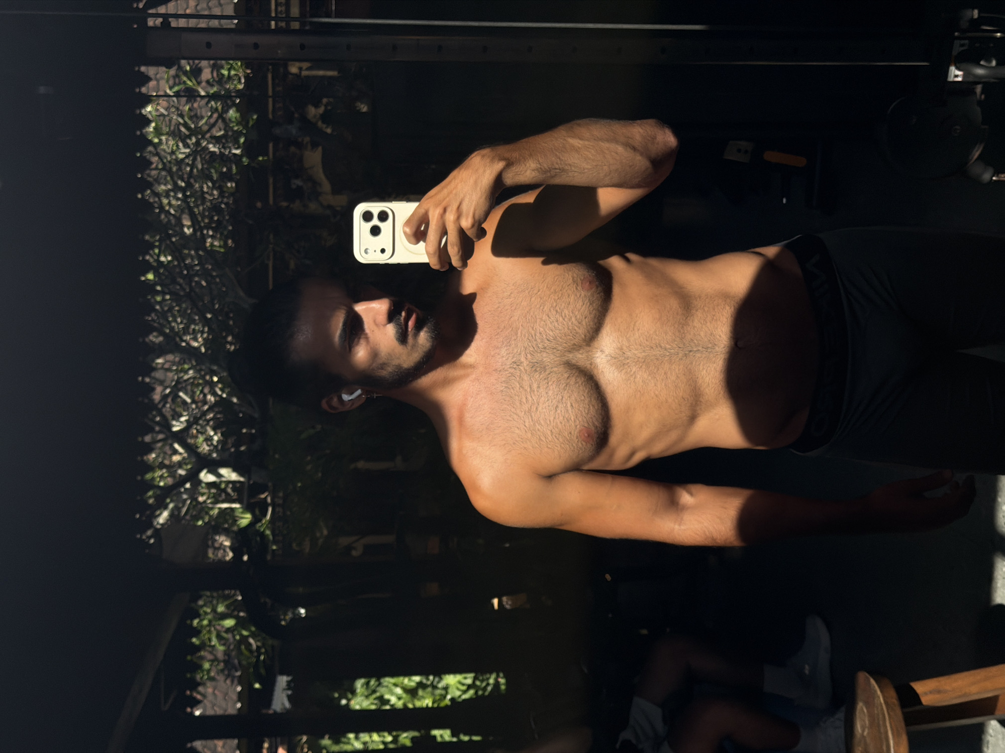 A shirtless man is holding a mobile phone in his right hand and taking a selfie in front of a reflective surface, likely a mirror. The man has short, dark hair and a trimmed beard. He is wearing dark-colored athletic shorts with a visible waistband. He appears to be in a gym setting, with a partial view of weightlifting equipment in the background. Reflected in the mirror is outdoor vegetation, suggesting the gym is near an open area. The image captures a moment of self-documentation, likely after or during a workout. It is in the city of North Kuta, Indonesia.