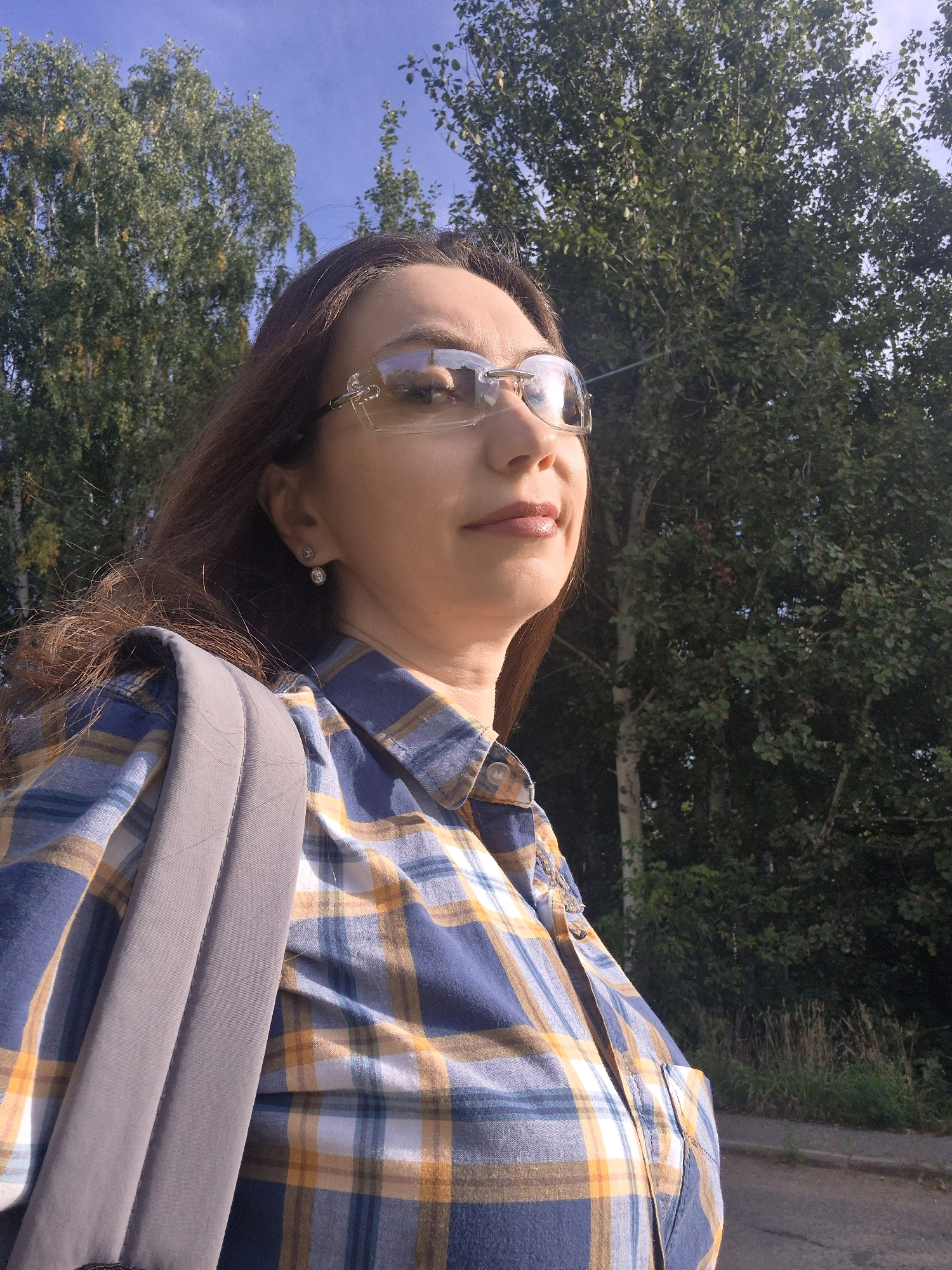 A woman with brown hair and wearing glasses and a plaid shirt is pictured outdoors with trees and a blue sky in the background. A backpack strap is visible over her shoulder. The scene appears to be a road or path next to a grassy area with trees. The location is Chaikovsky, Russia.