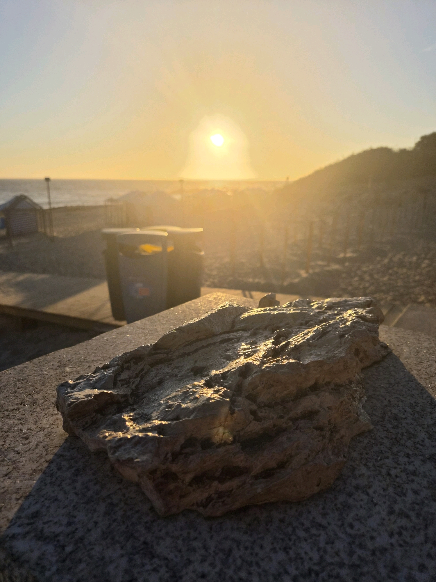 This is a close-up, low-angle shot of a rock in the foreground, bathed in the warm, golden light of a setting sun. The rock is rough and textured, with highlights glinting off its wet surface, suggesting it may have been recently rained on or is located near water. It rests on a speckled grey surface, possibly a stone wall or ledge.
In the mid-ground, a grey trash bin with a yellow lid is visible. Beyond the bin, the scene transitions to a beach environment. The sand is visible, with some scattered debris or possibly beach grass. A small wooden structure, resembling a beach hut or changing room, is partially visible on the left. Faintly in the background, there are more beach structures and a low, dark hill covered in vegetation, silhouetted against the bright sky.
The sky is a soft gradient of pale blue at the top, transitioning to a warm yellow and orange as it nears the horizon, where the sun is a bright, diffused orb. The sunlight casts a haze over the beach scene, creating a slightly blurred, atmospheric effect, especially in the distance. The presence of the ocean is indicated by the horizontal line of the water at the far horizon. The overall impression is a tranquil beach scene at sunset, with a focus on the textural details of the rock in the foreground.
The prompt asks to include the location in the city of Esposende, Portugal. However, there are no specific landmarks or visual cues within the image that definitively identify it as Esposende, Portugal.