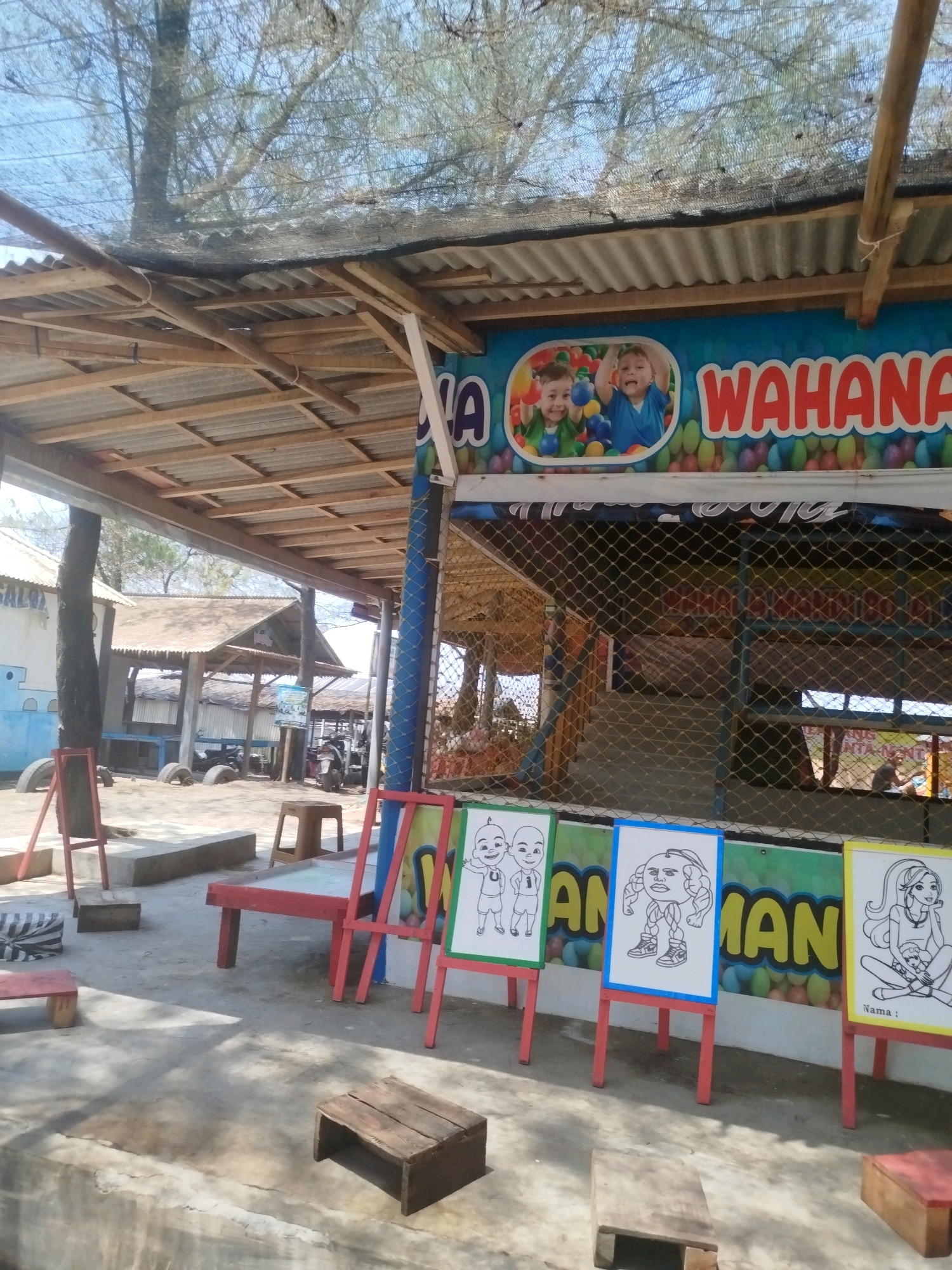 The image shows an outdoor play area with a covered structure, possibly part of an amusement facility, located in Setrojenar, Indonesia. The structure has a corrugated metal roof supported by wooden beams. A brightly colored banner with the word "WAHANA" and images of children playing in a ball pit is visible above the entrance. In front of the structure, three easels stand, each displaying a large drawing for children to color. The drawings depict cartoon characters. The ground is paved with concrete, and several small wooden stools and a red bench are scattered around the area. In the background, there are other simple structures, trees, and a motorcycle. The overall impression is that of a casual, child-friendly outdoor entertainment venue.