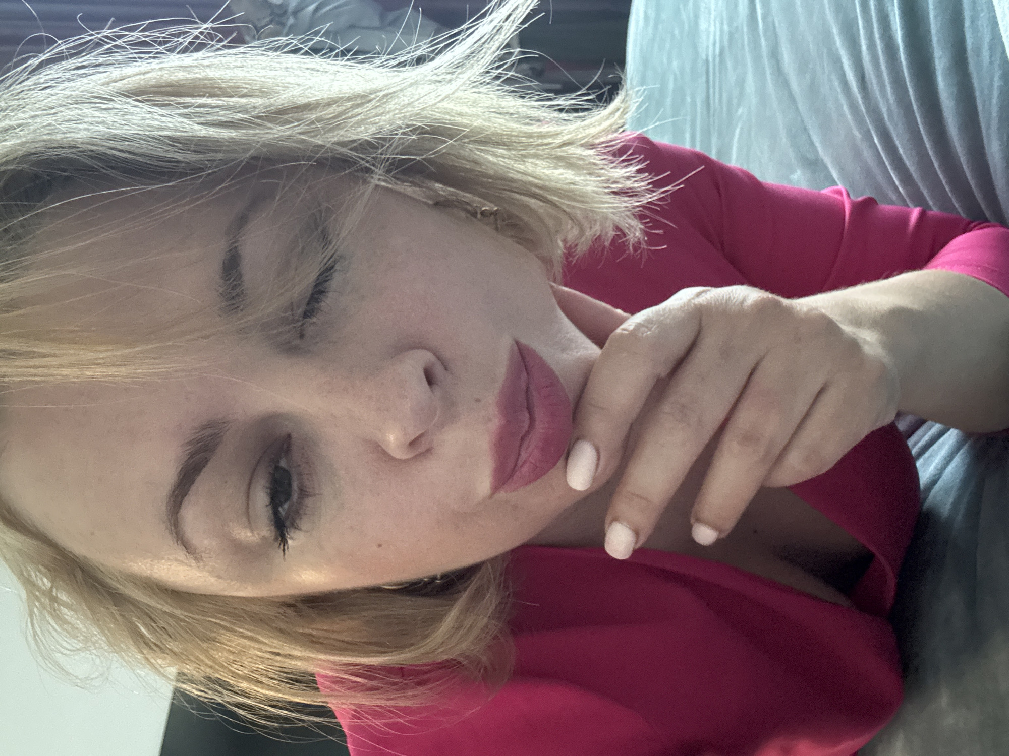 A blonde woman in a bright pink top is lying down on a bed. Her left eye is closed, and her right eye is open. Her hand is resting on her chin. The room appears to be a bedroom with a grey bedspread and what looks like curtains in the background. There is no indication of the city of Sochi, Russia, in the image.