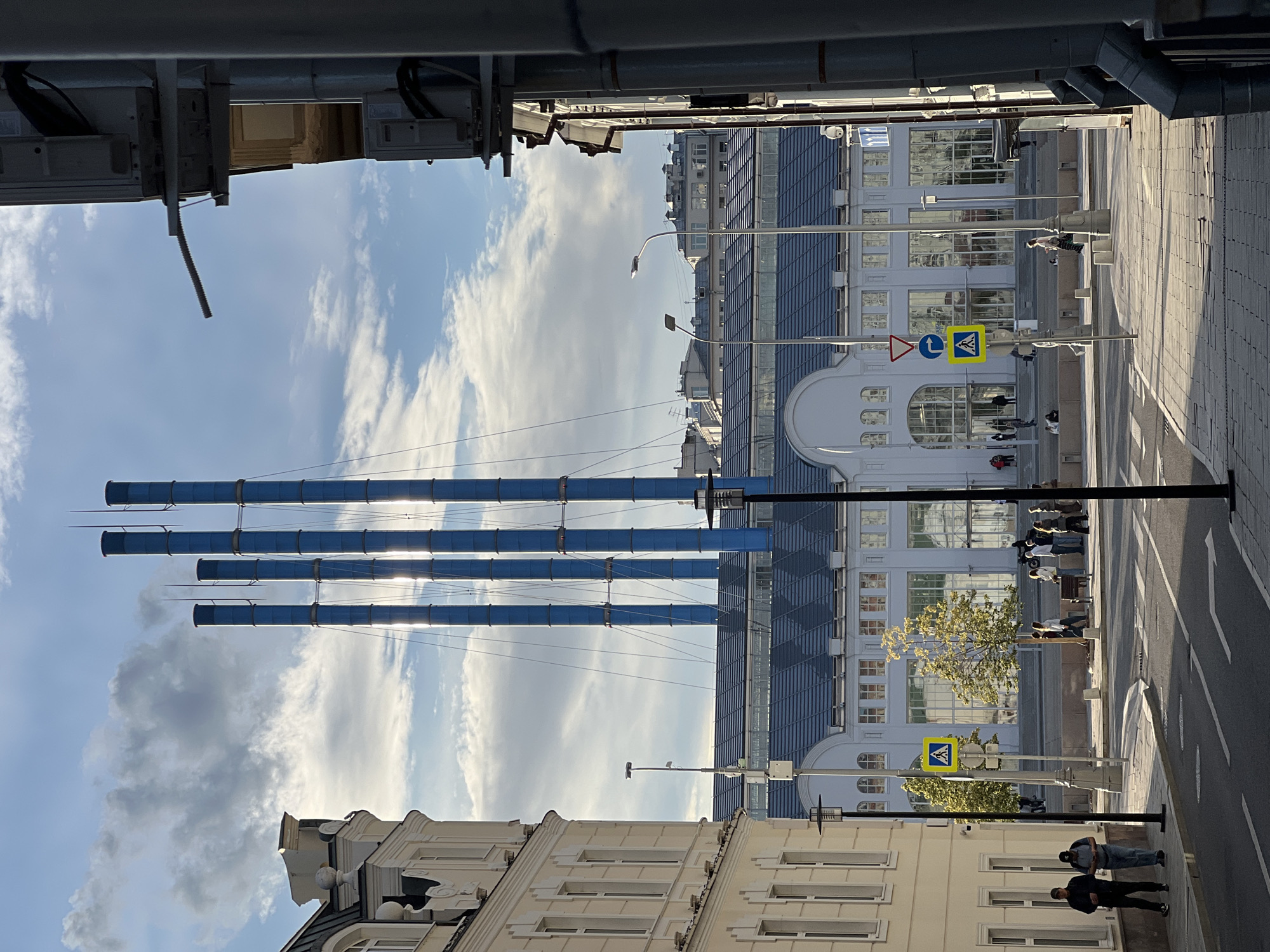 This image depicts a street scene in Moscow, Russia, bathed in the warm, low light of late afternoon or early morning. The sun casts long shadows across the paved sidewalk and roadway, suggesting a clear day with a few scattered clouds in the bright blue sky. Dominating the upper left portion of the frame are four prominent, vertical blue pipes, possibly part of an industrial installation or artistic structure.
In the background, modern and older buildings line the street. One building features a contemporary facade with a dark, reflective glass exterior, punctuated by numerous windows. Adjacent to it, a lighter-colored building displays classic architectural elements, including arched windows and decorative moldings.
Several people are visible on the sidewalk, some walking, others standing or sitting. Their presence suggests a normal urban activity, perhaps commuting or enjoying the outdoors. One group of people is gathered near a crosswalk, indicated by a yellow pedestrian crossing sign. Another individual is seen walking down the sidewalk towards the viewer.
The overall atmosphere is one of a bustling yet serene urban environment. The lighting creates a slightly dramatic and picturesque quality, highlighting the architectural details and casting a warm glow on the scene. No visible text is readily apparent in the image that provides specific location or informational context.