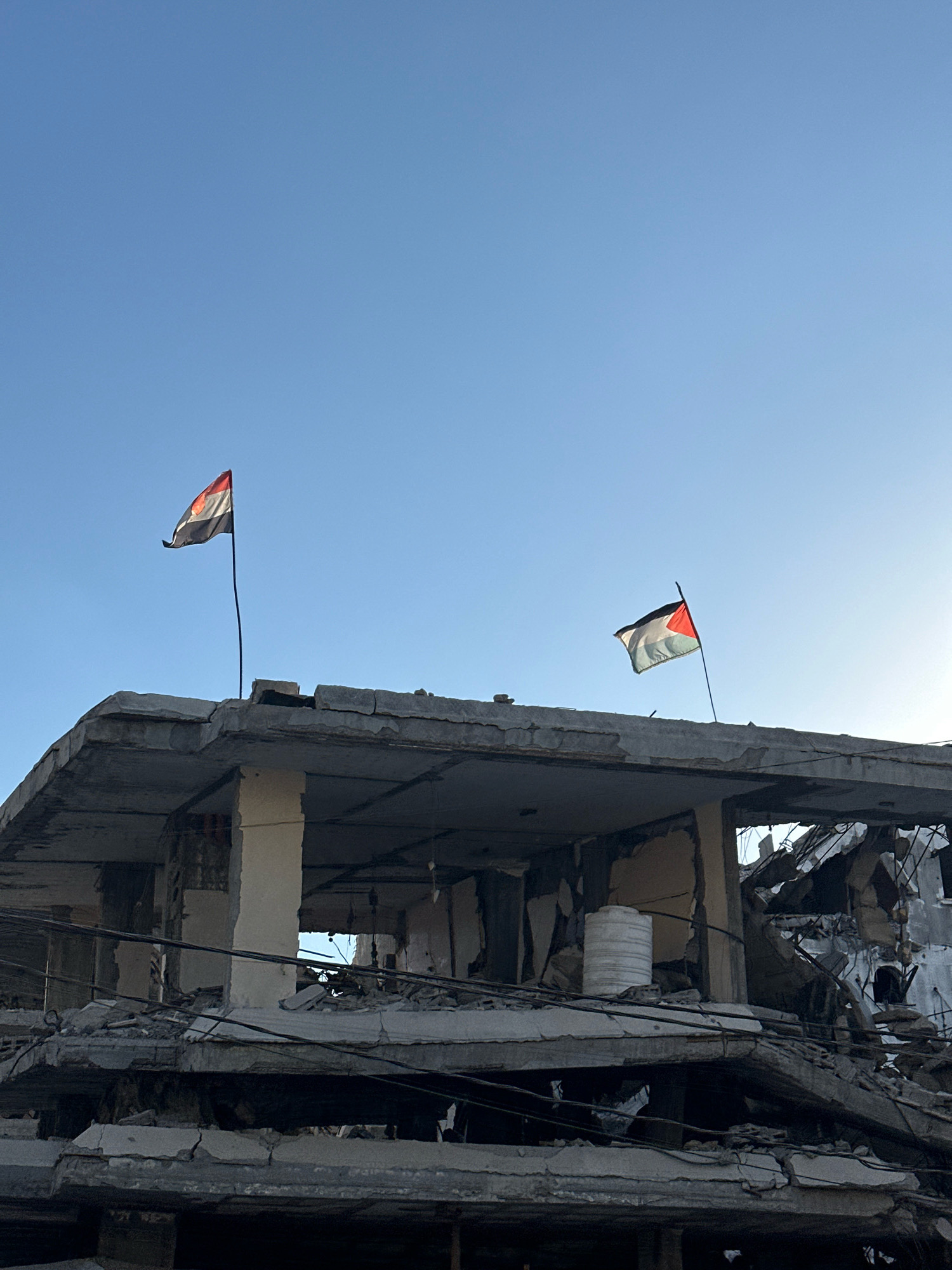 Two flags are visible against a clear blue sky, flown from the rooftop of a severely damaged building. The building appears to be a multi-story structure, with sections of its concrete floors and walls collapsed and debris scattered across its surfaces. The flag on the left is partially obscured and appears to be a horizontal tricolor with red, white, and black bands, possibly the flag of Syria. The flag on the right is clearly visible as the Palestinian flag, with a black, white, and green horizontal tricolor and a red triangle at the hoist. Wires are visible hanging from the damaged structure. The scene depicts a post-conflict environment within Gaza, Palestinian Territories, with evidence of destruction.