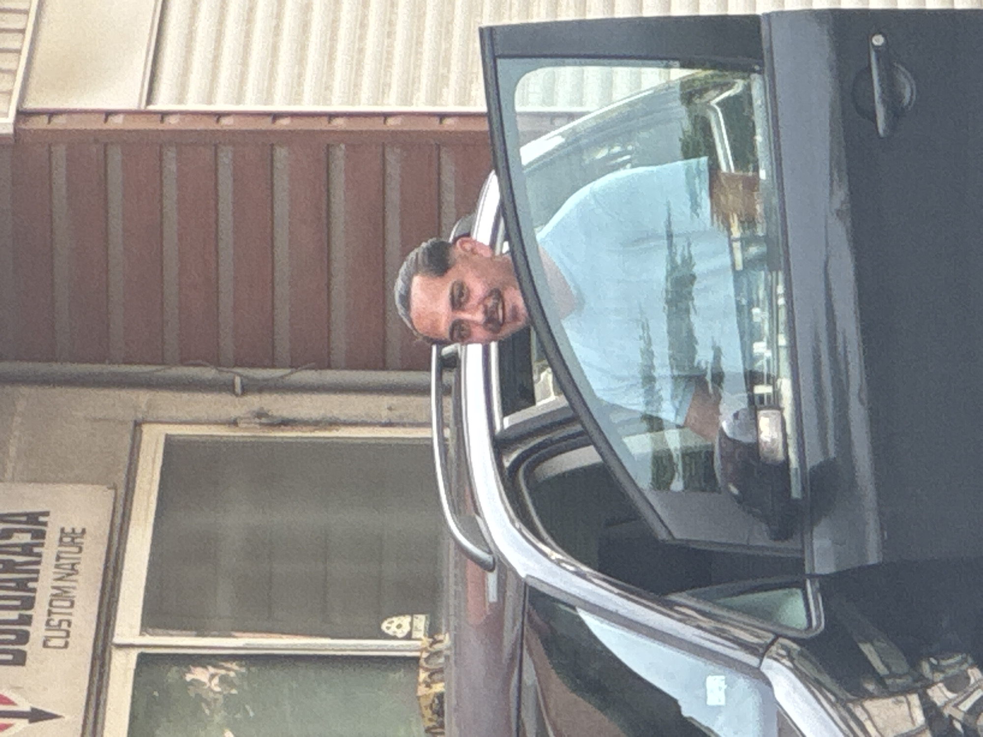 A cheerful man leans out of the open driver's side door of a dark-colored vehicle, likely an SUV or truck. He is middle-aged, with dark hair, a mustache, and a goatee, wearing a light blue t-shirt. The man smiles broadly directly at the camera, conveying happiness and friendliness. The car features chrome trim along the window frame and an extended side mirror.
In the background, a building with reddish-brown vertical siding on its upper portion and a lighter, possibly corrugated metal, roof is visible. Below the siding, another section of the building features a large window or glass panel. A partially visible white sign on the building to the lower left reads "...GULARASA CUSTOM NATURE," likely indicating "BULGARASA" as the main text.
The scene takes place during the daytime under bright, sunny conditions, suggested by the clear lighting and reflections. The overall setting appears to be an urban or commercial area, consistent with a location such as Varna, Bulgaria.