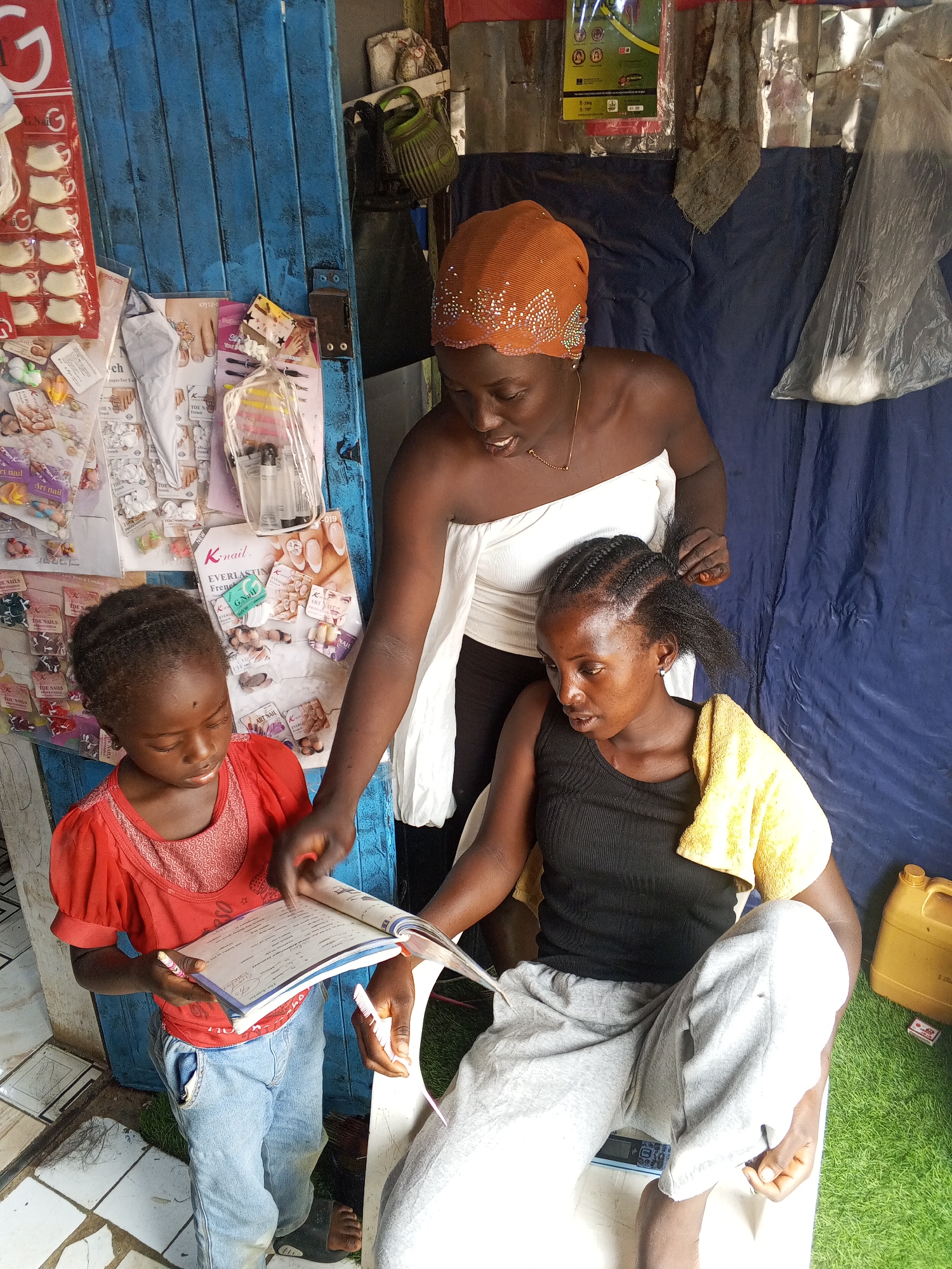 A photograph depicts three African women; one woman is seated having her hair braided by another, while a young girl stands next to them reading a book. The setting appears to be a makeshift hair salon in Gwong, Nigeria, judging by the nail art advertisements affixed to a blue door, the blue draped cloth backdrop, and the artificial grass flooring. The seated woman is wearing a black tank top and grey sweatpants, with a yellow towel draped over her shoulder. The woman braiding her hair is wearing an orange headscarf, a white, off-the-shoulder top, and dark pants. The girl is wearing a red shirt and blue jeans. All three are closely engaged with each other, with the girl pointing at something in the book for the seated woman. The environment is cluttered with various items, including a yellow jerrycan, a box, and plastic-wrapped items hanging in the background.