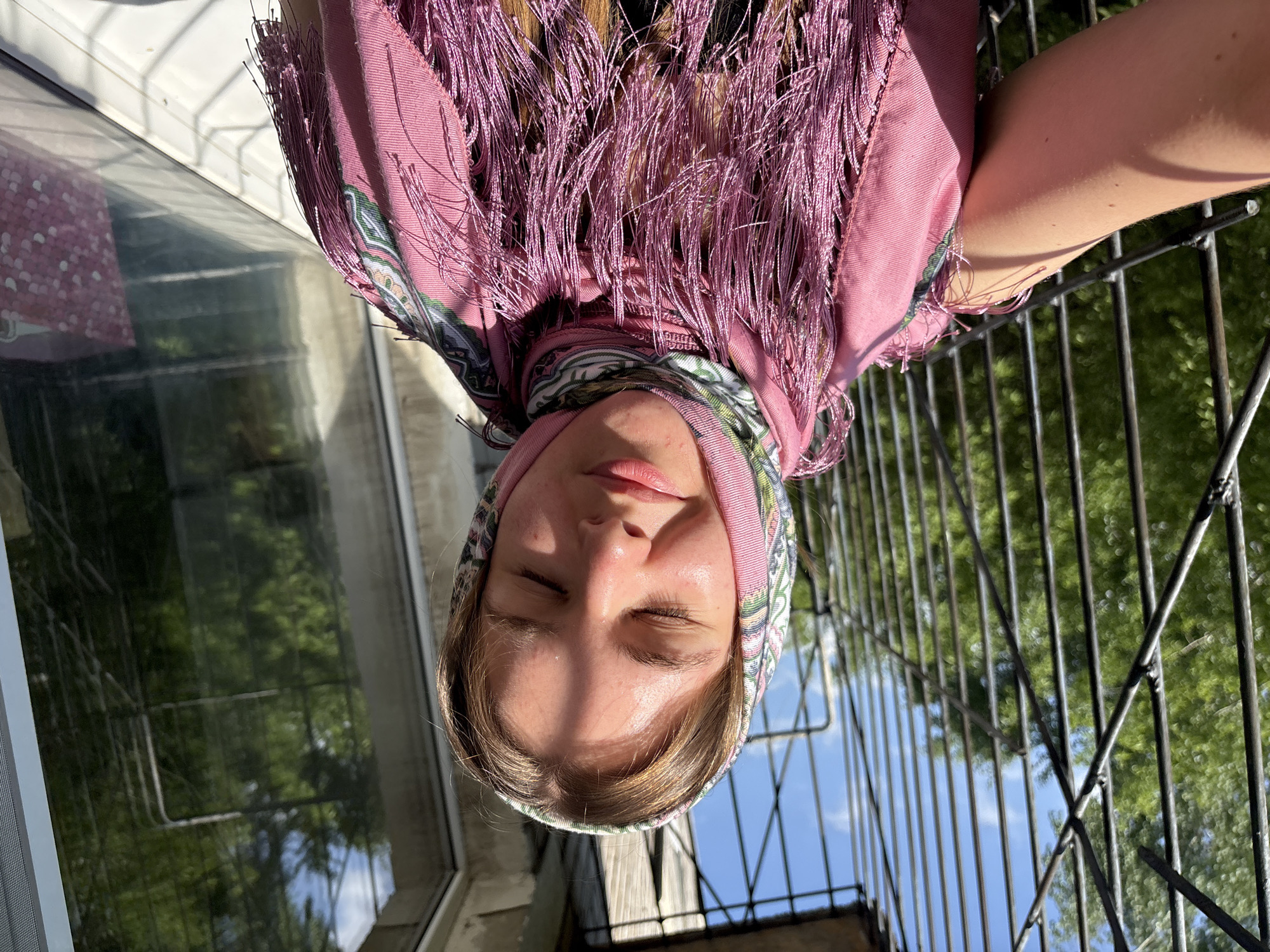 A young woman with closed eyes is wearing a pink headscarf with fringed edges. She is standing near a window and a black metal railing. The image was taken in Perm, Russia, as determined by the subject's headscarf, which is a traditional Orenburg shawl. The railing and window suggest she is likely on a balcony or near a building exterior. Trees and sky are visible in the background.
