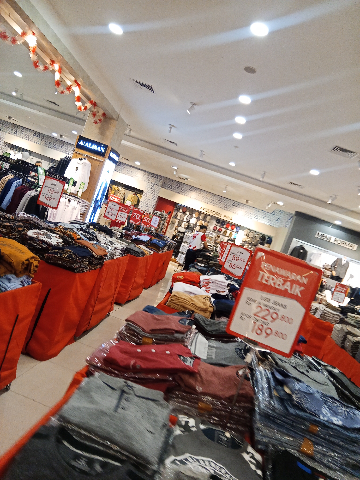 The image depicts the interior of a clothing store, likely a department store or a large boutique, located in Lubuklinggau, Indonesia. The store is well-lit with overhead fluorescent lights and appears to be actively displaying merchandise for sale, with a focus on men's clothing. Several racks of shirts and other apparel are visible, with some items neatly folded on display tables covered in orange fabric. Red and white decorative garlands are strung across the ceiling, suggesting a festive or promotional period. Prominent red and white signage with Indonesian text, including pricing and promotional offers such as "PENAWANAN TERBAIK" (Best Offer) and specific price reductions, are displayed, indicating a sale event. Some prices are listed in Indonesian Rupiah (Rp). The aisles are tiled, and the overall environment suggests a retail setting aimed at selling fashionable clothing. The image is taken from a slightly low, angled perspective, giving a dynamic feel to the scene.