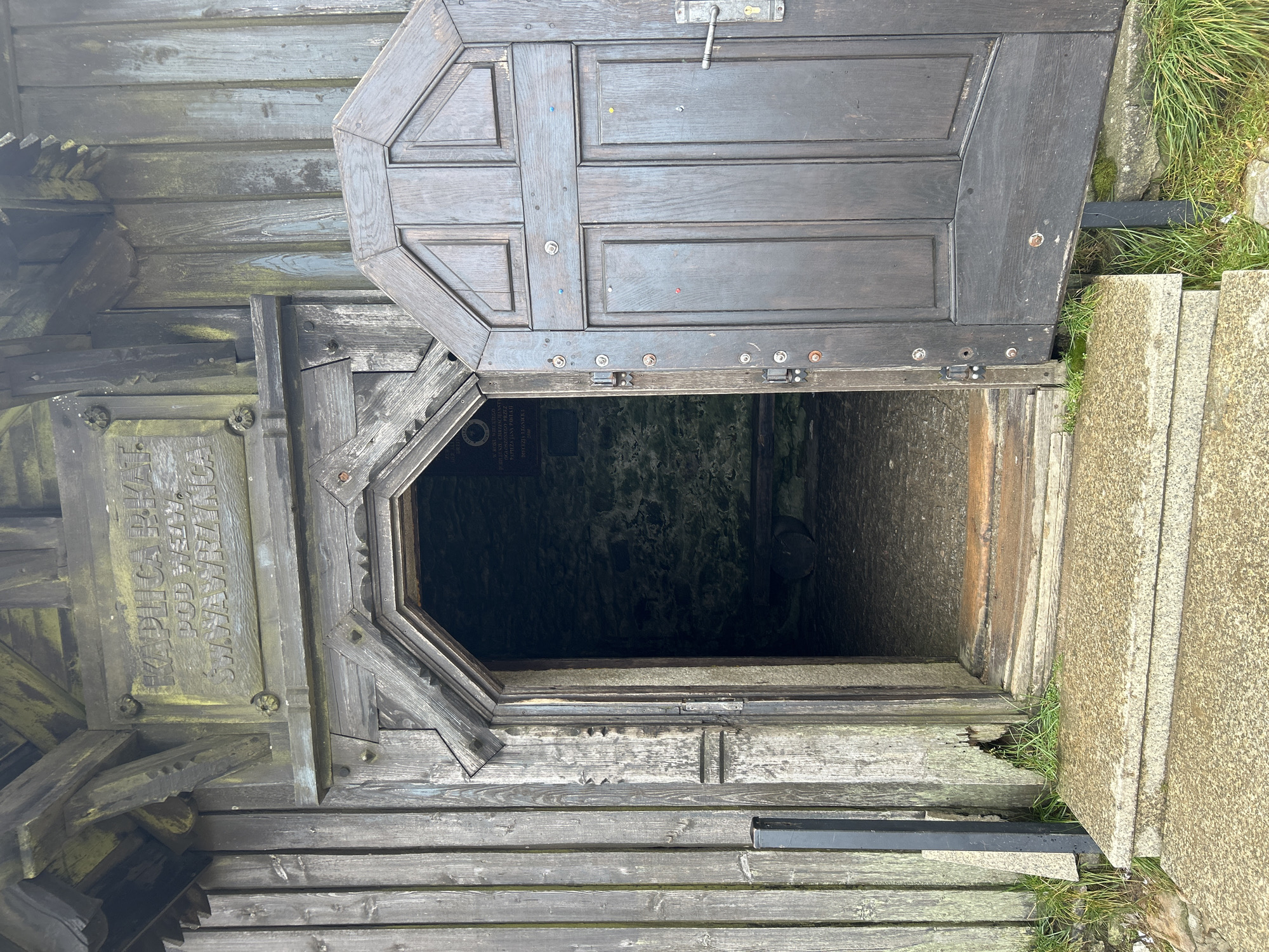 The image shows the entrance to a small wooden building, possibly a chapel or shelter, situated in Malá Úpa, Czechia. The building is constructed of weathered, dark wood with a mossy patina, suggesting it is old and exposed to the elements. Above the entrance, a wooden plaque is affixed to the facade with carved lettering. The main entrance is an open, arched doorway with a dark wooden door ajar to the right. The door has multiple panels and is secured with a metal handle and latch. Colorful thumbtacks or small nails are scattered across the surface of the open door. Inside the dimly lit building, a stone wall is visible, and a rough-hewn wooden bench or seat can be seen. Steps made of rough-hewn stone lead up to the entrance. The surrounding environment appears to be natural, with grass growing around the base of the structure. The overall impression is one of rustic simplicity and historical character.