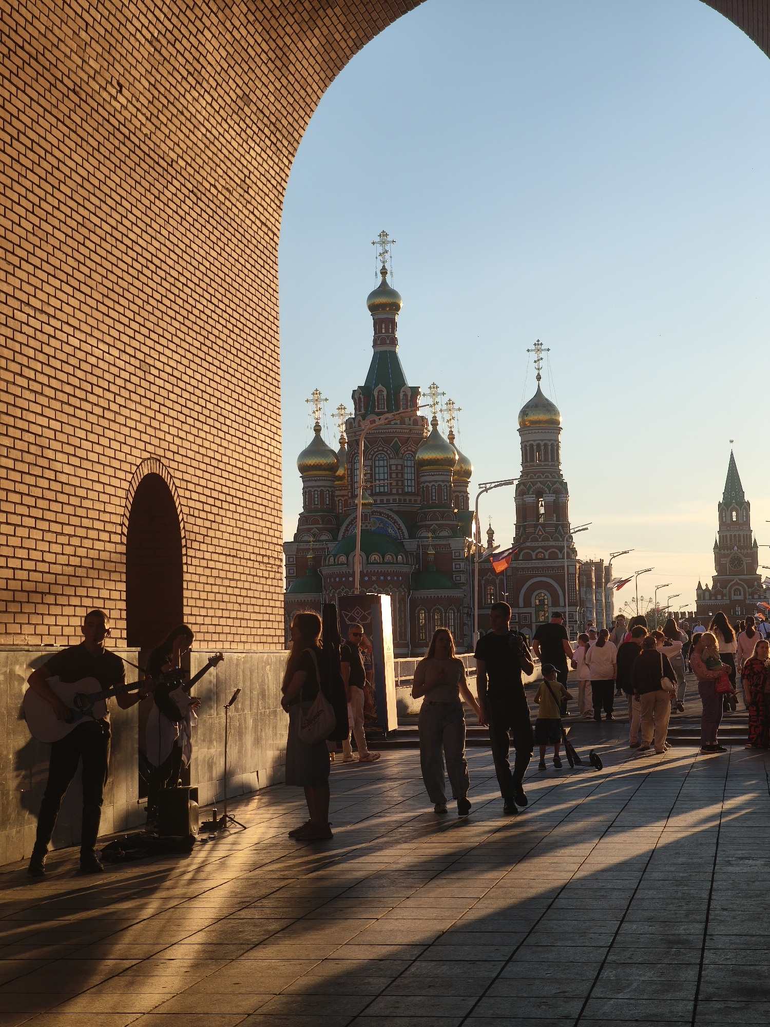 The image depicts a sunny scene in Yoshkar-Ola, Russia, possibly on Yoshkar-Ola National Congress Square. In the foreground, an arched brick structure frames a view of a street lined with people. On the left, under the arch, a musical trio is performing, with two individuals playing guitars and another holding one. They are positioned near a brick wall with an arched alcove. A portable speaker sits on the ground near them.
Further into the scene, a crowd of people walks along a paved walkway. In the background, the distinctive onion domes and spires of several Orthodox churches, specifically the Annunciation Cathedral and the Tsar Kokhno Cathedral, dominate the skyline. The architecture is ornate with vibrant colors, and crosses adorn the tops of the domes. Russian flags are visible on poles. The sunlight casts long shadows across the pavement, indicating it is late afternoon or early evening. The sky is a clear, pale blue, with no clouds visible.