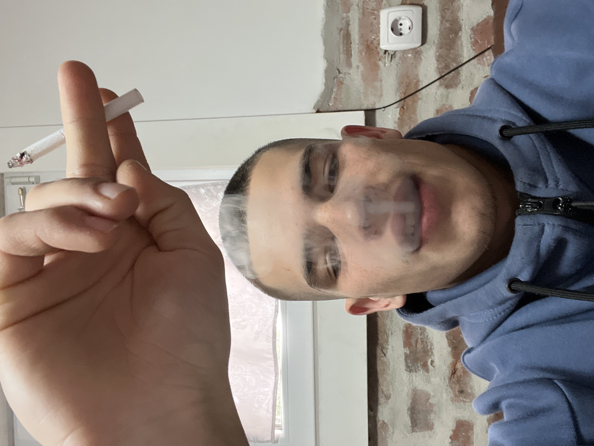 A man wearing a blue hoodie is holding a lit cigarette between his fingers. He appears to be exhaling smoke. The background includes a window, a white wall, and an exposed brick wall with a white electrical outlet. The location in Plovdiv, Bulgaria cannot be determined from this image.