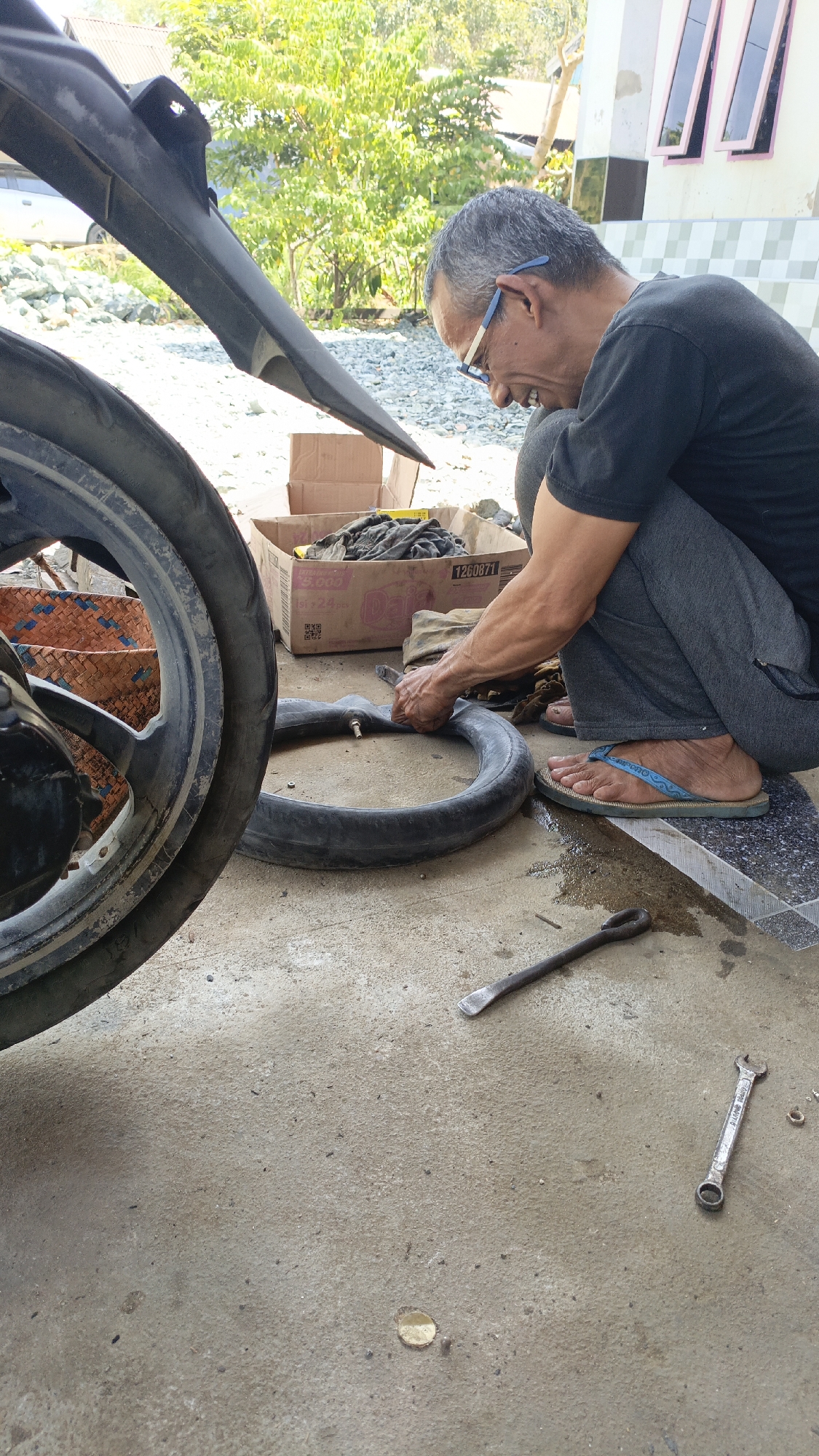 An older man with graying hair and glasses is seen squatting on a concrete ground, engaged in repairing a motorcycle inner tube. He is wearing a dark t-shirt and gray pants, with blue flip-flops on his feet. In front of him lies the detached inner tube of a motorcycle tire, which he is manipulating with his hands. Nearby, a toolbox or box filled with rags is visible, along with tools like a tire lever and a wrench scattered on the ground. The scene appears to be an outdoor setting, possibly a yard or roadside in the city of Saring Sungai Binjai, Indonesia. In the background, a house with windows and some greenery are visible, suggesting a residential area. The overall atmosphere is that of a practical, everyday repair task being carried out in a simple, outdoor environment.