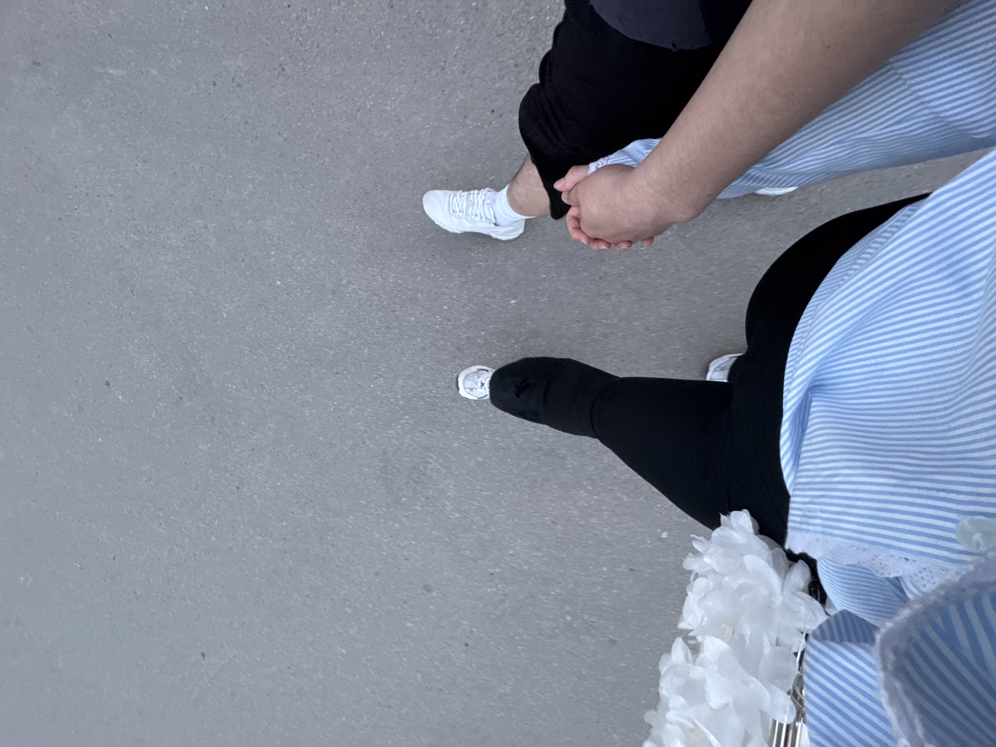 Two individuals are walking on a grey paved surface, likely a street or sidewalk. One person is wearing black pants and white sneakers. The other person is wearing black leggings, white sneakers, and a blue and white striped shirt. They are holding white decorative flowers. The location is Perm, Russia.