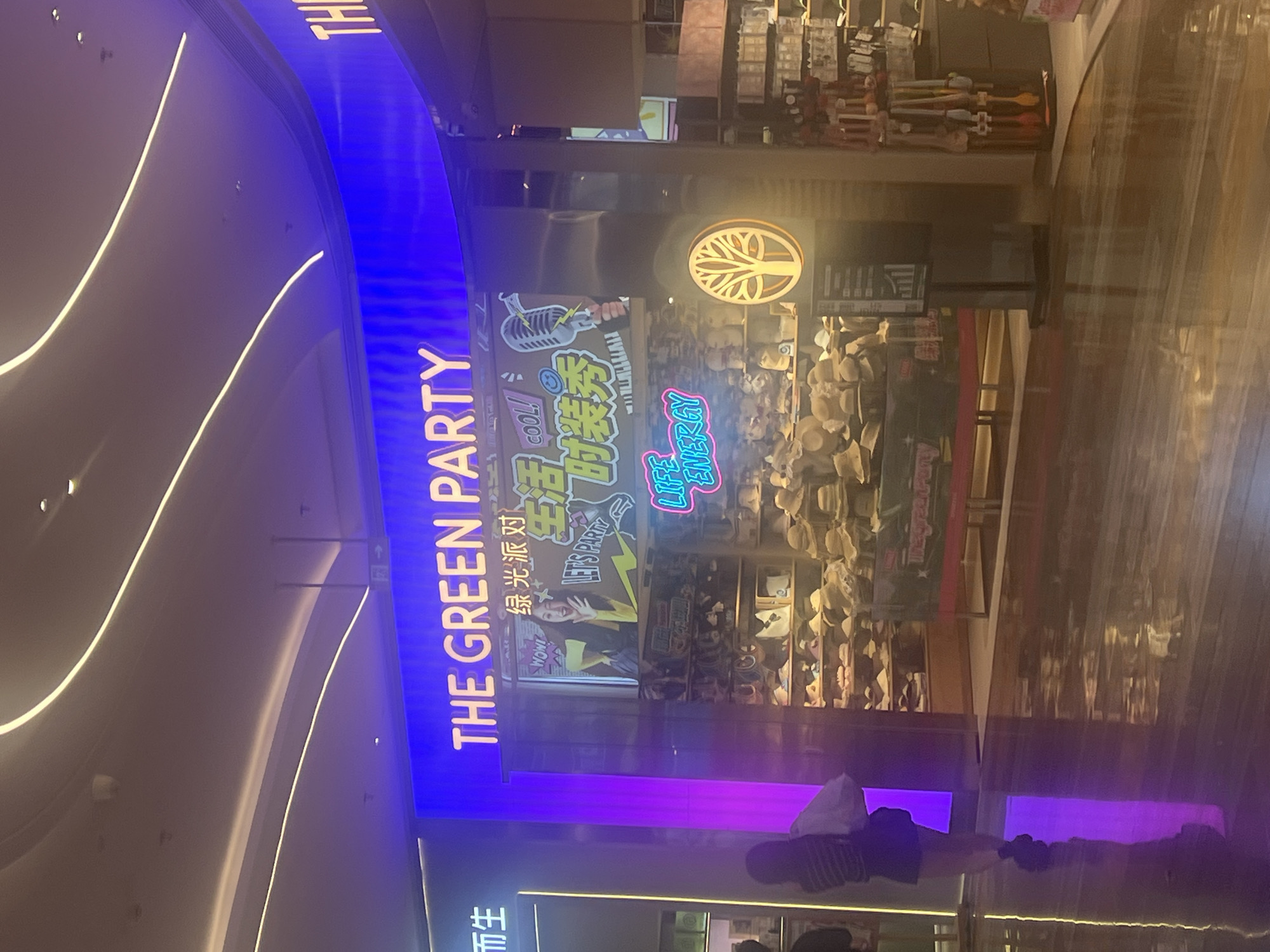 A storefront named "THE GREEN PARTY" is brightly lit with blue neon lights. Above the entrance, the store's name is emblazoned in large white letters. Below this, a digital screen displays an image of a woman holding a microphone, with Chinese text and cartoon elements suggesting a fashion show or entertainment event. The store's interior showcases shelves filled with hats, likely for sale, and other merchandise. A stylized tree logo is visible on the right side of the storefront. The floor is highly reflective, mirroring the lights and store facade. In the foreground, a person is walking away from the camera, dressed in casual clothing and carrying a white bag. The environment appears to be an indoor shopping mall. This scene is located in Liandu District, China.