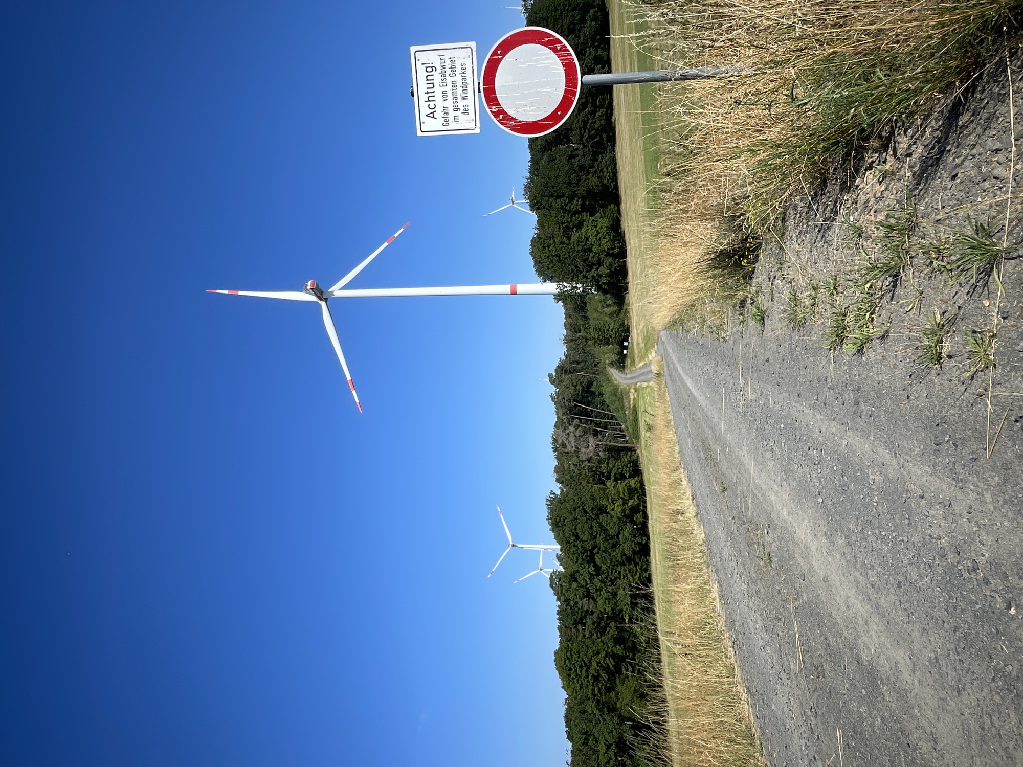 In the city of Berfa, Germany, a gravel road winds through a grassy field towards a wind farm under a clear blue sky. In the foreground, dry grass lines the side of the road. To the right, a signpost displays a warning message in German, reading "Achtung! Gefahr von Eisabwurf im gesamten Gebiet des Windparkes" (Attention! Danger of ice falling in the entire area of the wind farm), along with a round, red and white prohibitory sign. Several wind turbines are visible in the middle ground and background, their white towers and red-tipped blades stark against the sky. The turbines are positioned amidst a line of dense green trees. The scene depicts a rural landscape dominated by renewable energy infrastructure.