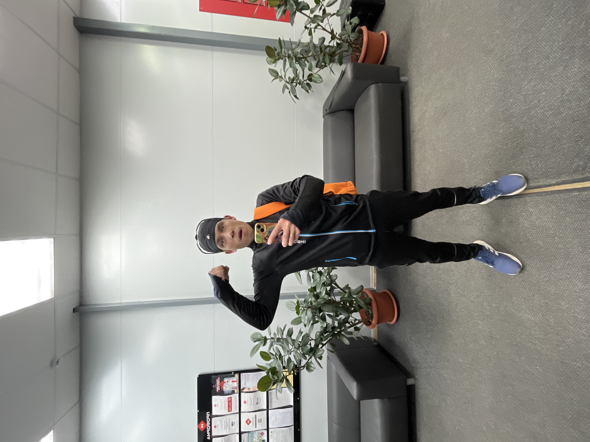 A man stands in a room, flexing his arm. He is wearing a black tracksuit with blue stripes, a black baseball cap, and blue running shoes. An orange bag is slung across his chest. He is holding a cell phone in his other hand.
Two grey couches are in the room. A potted plant sits on each of the couches. A board with documentation is affixed to the wall.
The location is Chaikovsky, Russia.