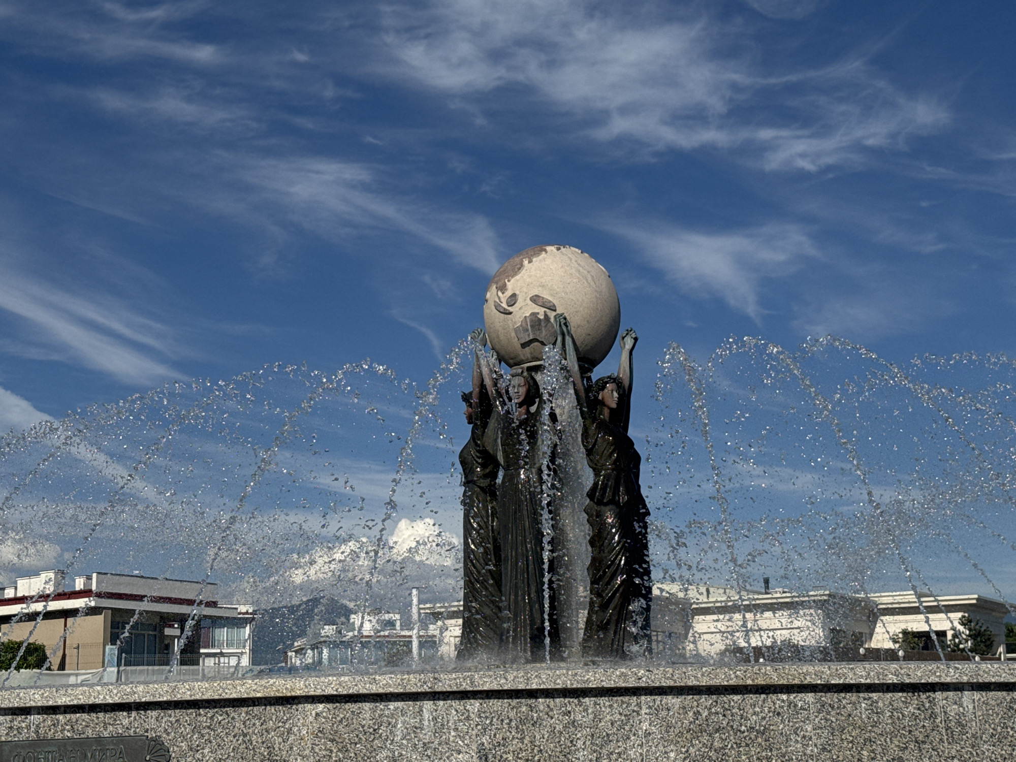 The image displays a prominent fountain featuring a central sculpture under a clear, bright blue sky adorned with scattered wispy clouds. The sculpture depicts three dark-colored female figures, likely made of bronze, standing in a circle and holding aloft a large, light-colored sphere that appears to be a globe representing the Earth, complete with sculpted landmasses.
Water jets from the fountain create a broad, arching spray that cascades over and around the figures, giving them a glistening, wet appearance. The strong sunlight causes numerous sparkling reflections on the water droplets.
In the background, visible through the transparent sheets of water, are several modern buildings with light-colored walls and reddish roofs. Faint outlines of distant mountains or hills can be seen beyond these structures. The base of the fountain is constructed from speckled granite. An inscription on the fountain's base in Cyrillic script reads "ФОНТАНЫ МИРА" (Fontany Mira), which translates to "Fountains of Peace" or "Fountains of the World," alongside a shell emblem. The scene takes place during bright daylight in an outdoor public space, specifically located in Sirius, Russia. The primary activity is the continuous operation of the fountain.
