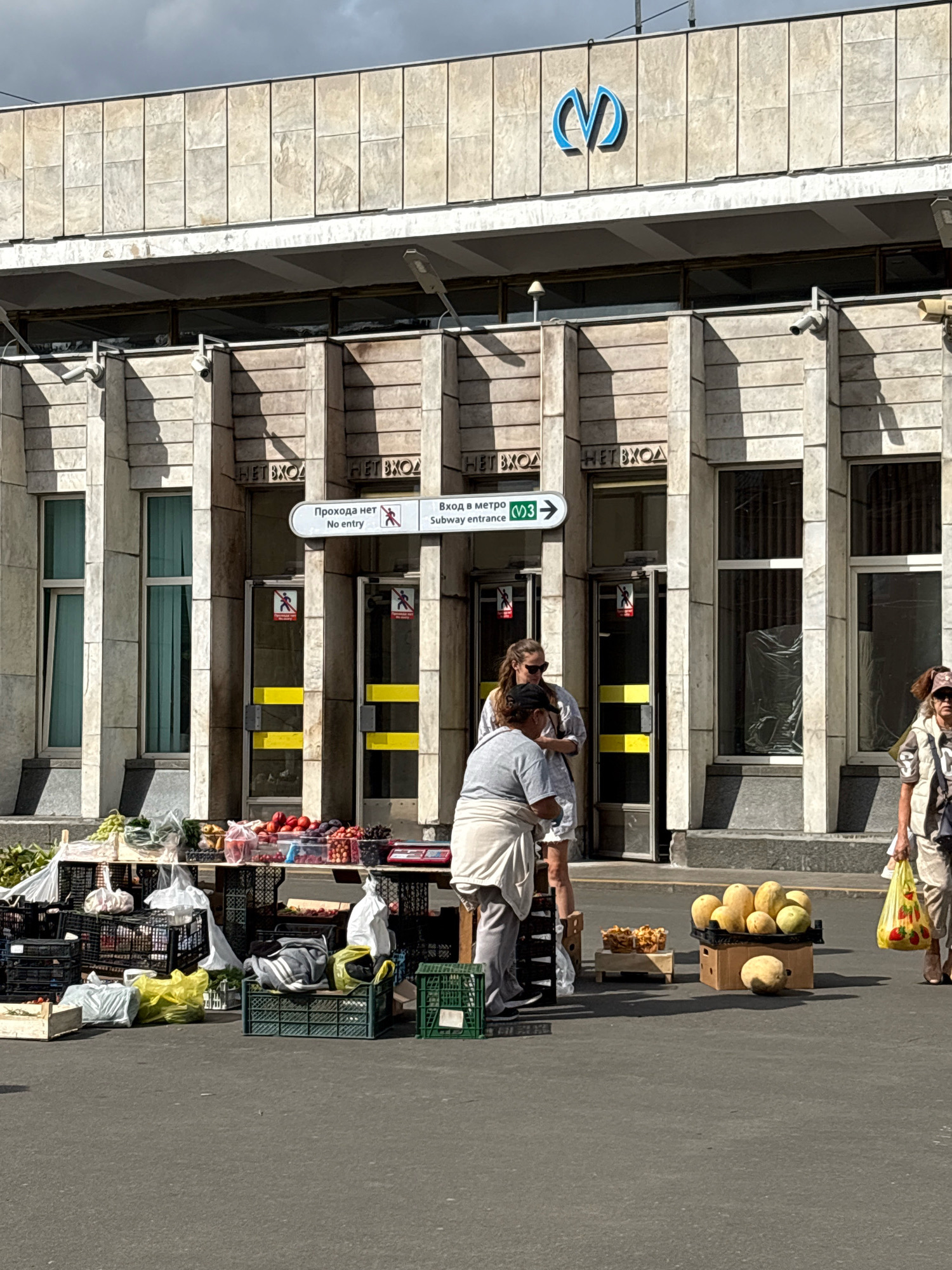 The image captures an outdoor scene in front of a building with a facade of light-colored stone panels. A blue metro logo is visible at the top center of the building. Below the logo, there are entrances to the metro, with signs indicating "No entry" in Russian and English, and a directional sign pointing towards "Subway entrance M3".
In the foreground, a street market is set up. Several crates and tables are laden with a variety of fruits and vegetables, including grapes, plums, apples, cherries, melons, and what appear to be some baked goods. Two women are present at the market. One woman, wearing a grey t-shirt and a white apron, is standing behind a stall, possibly tending to the produce. Another woman, in a white shirt and sunglasses, is standing next to her. A third person, partially visible on the right side of the image, is holding a yellow plastic bag filled with produce and is walking away from the market. The ground is paved, and the lighting suggests it is daytime with bright sunlight casting shadows.