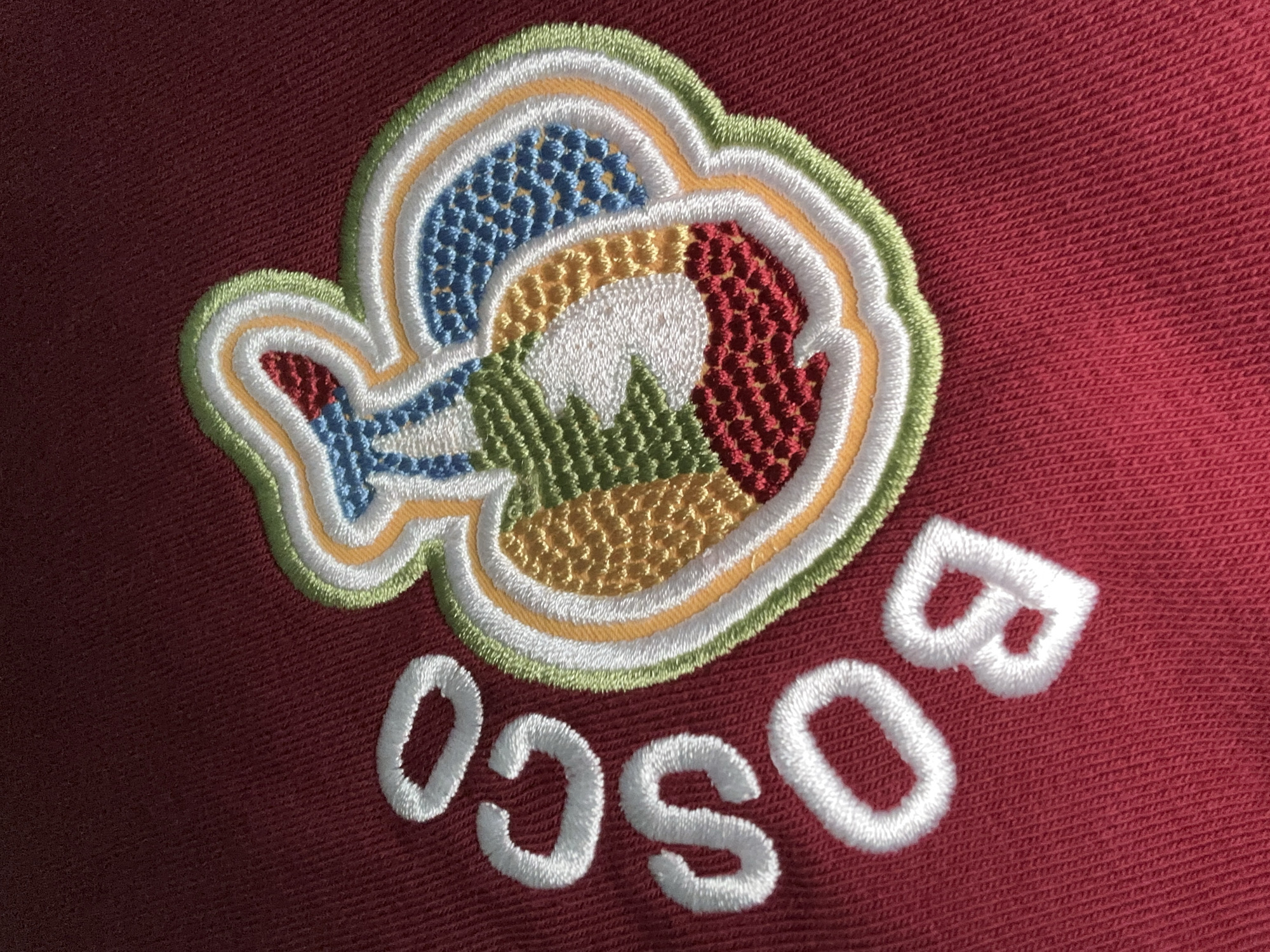 The image shows a close-up of an embroidered patch on a maroon-colored fabric. The patch is cloud-shaped and contains multiple colors: blue, white, red, yellow, and green. Below the patch, the word "BOSCO" is embroidered in white letters, in a curved pattern. The location in the city of Baranovka, Russia, is not discernible from the image.