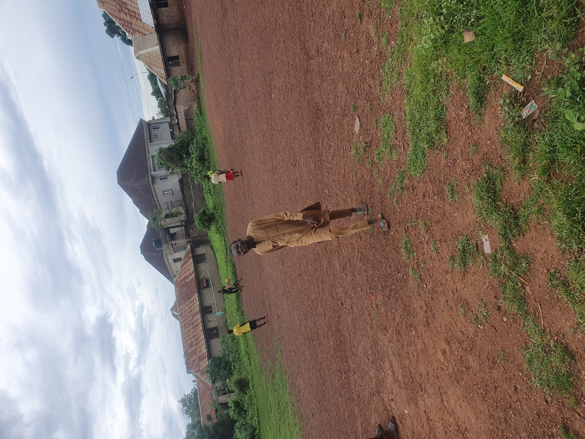 This image, captured in Idah, Nigeria, depicts an outdoor scene on a reddish-brown dirt field with interspersed patches of green grass. Four individuals are present, engaging in recreational activities, against a backdrop of various residential structures under an overcast sky.
In the foreground, slightly to the right of the center, a dark-skinned male subject is walking towards the viewer, smiling directly at the camera. He wears a light brown or tan long-sleeved tunic and matching trousers, with light blue sandals on his feet. His left hand is partially visible, holding a dark, rectangular object against his thigh.
In the mid-ground, three additional male subjects are visible. To the left, a subject in a yellow t-shirt and dark shorts stands facing slightly away from the camera. In the center-right, a subject in a dark t-shirt and dark shorts is actively engaged with a black and white football (soccer ball) at his feet, suggesting play. Further to the right and slightly behind, a fourth subject in a red top and dark shorts is also on the field, partially obscured, and appears to be involved in the same activity. A makeshift net, possibly for volleyball or football, is stretched horizontally across the mid-background between two unidentified supports.
The ground consists of reddish-brown laterite soil, characteristic of the region, with numerous small pebbles and debris. Scattered clumps of green grass grow unevenly across the field. In the background, a series of buildings are visible. To the left, a single-story structure with a rusty corrugated metal roof and unpainted concrete block walls features multiple windows. In the center-background, a larger, two-story grey building with a dark-colored hip roof and white-framed windows is prominent. A water tank is mounted on a raised platform to the right of this building. Another structure with a rusty corrugated roof and unpainted walls is visible further to the right. Lush green trees and foliage are present throughout the background, situated behind and between the buildings. The sky is uniformly overcast with grey clouds. Litter, including a yellow wrapper and small white boxes, is visible on the ground in the foreground-right.