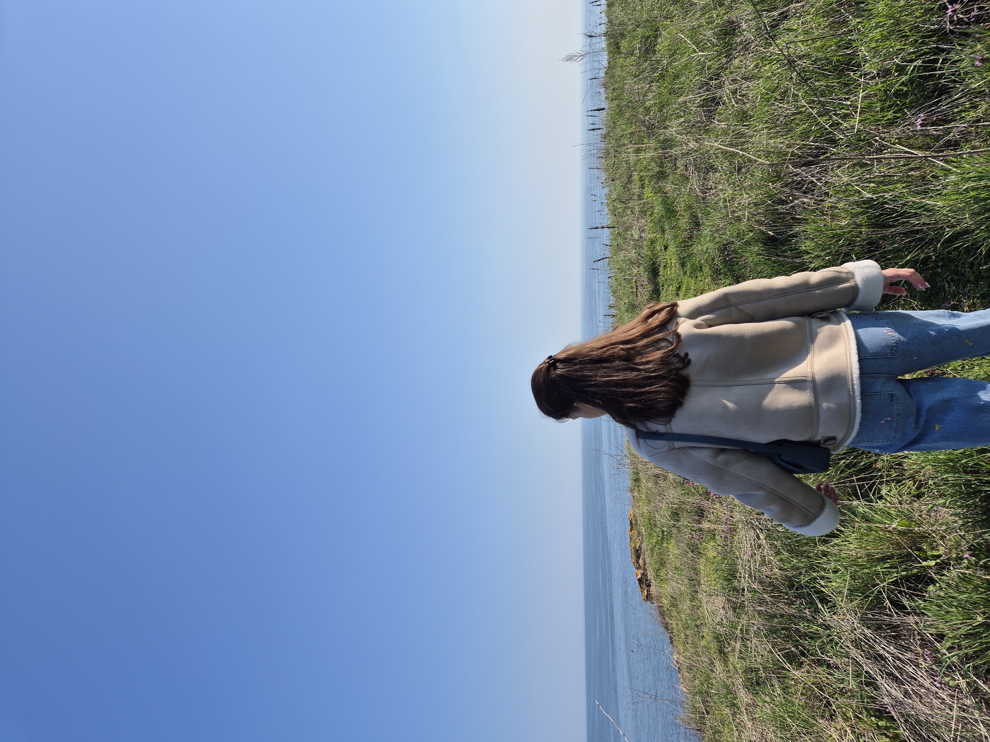 A woman stands on a grassy verge overlooking the Black Sea in Sveti Nikola, Bulgaria. She is wearing a beige shearling jacket, blue jeans, and has long brown hair. The sky is clear and blue. The scene is primarily composed of the sky, the sea, and the grassy verge.