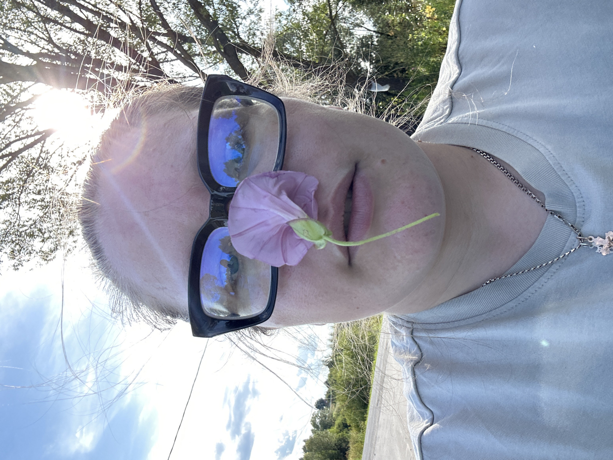 A close-up, low-angle shot captures a person wearing large black-framed glasses and a light gray t-shirt. A delicate pink flower is positioned directly in front of the person's nose, partially obscuring it. The background reveals a sun-drenched outdoor scene with a bright blue sky dotted with clouds. Sunlight streams through the branches of a tree on the right side of the frame, creating lens flares. A rural landscape with a road and rolling hills covered in greenery is visible in the distance, reflected in the person's glasses. The image appears to be taken in or near the city of Kalitsino, Russia, based on the visual cues of the landscape and vegetation.