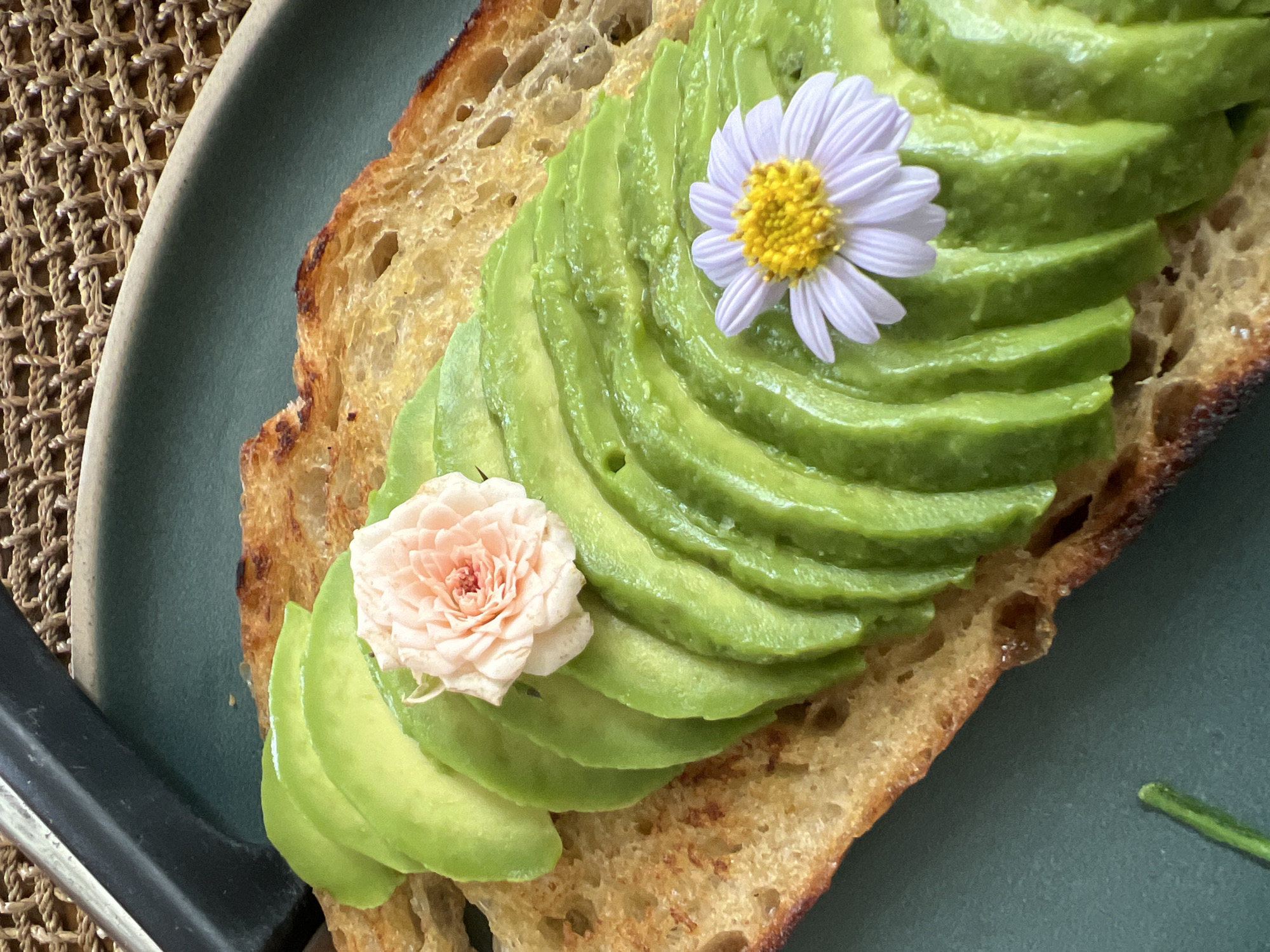A close-up, overhead shot shows a slice of toasted bread topped with fanned-out avocado slices. A delicate pink rosebud rests on the avocado near the top of the slice, and a small, white and yellow daisy is positioned further down. The toast is golden brown with some darker, toasted edges. The avocado is a vibrant green. The toast sits on a dark teal plate, which is placed on a woven placemat. The background is blurred, suggesting an outdoor or naturally lit setting. The image is cropped tightly, focusing on the food presentation. There are no people visible, and no specific location information can be determined from the visual content.