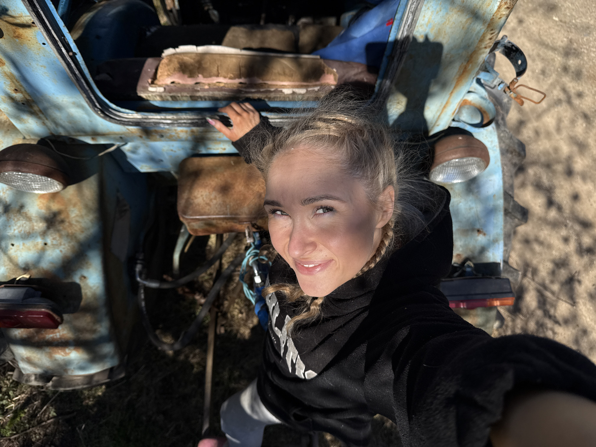 A fair-skinned woman with blonde hair styled in braids is the central subject of the image. She is wearing a dark-colored hooded sweatshirt with a visible design on the front. She appears to be smiling and looking up at the camera, which seems to be positioned from a lower angle, possibly a selfie perspective.
The woman is positioned in front of a dilapidated, light blue tractor with evident rust and wear. The tractor features large rear wheels and smaller front wheels, and its components are exposed due to its aged condition.
The setting appears to be outdoors, with ground cover consisting of grass, dry leaves, and some visible debris. It's daytime, with natural lighting casting shadows in certain areas of the scene.
No specific details connect this image to the city of Nizhnyaya Chermoda, Russia.