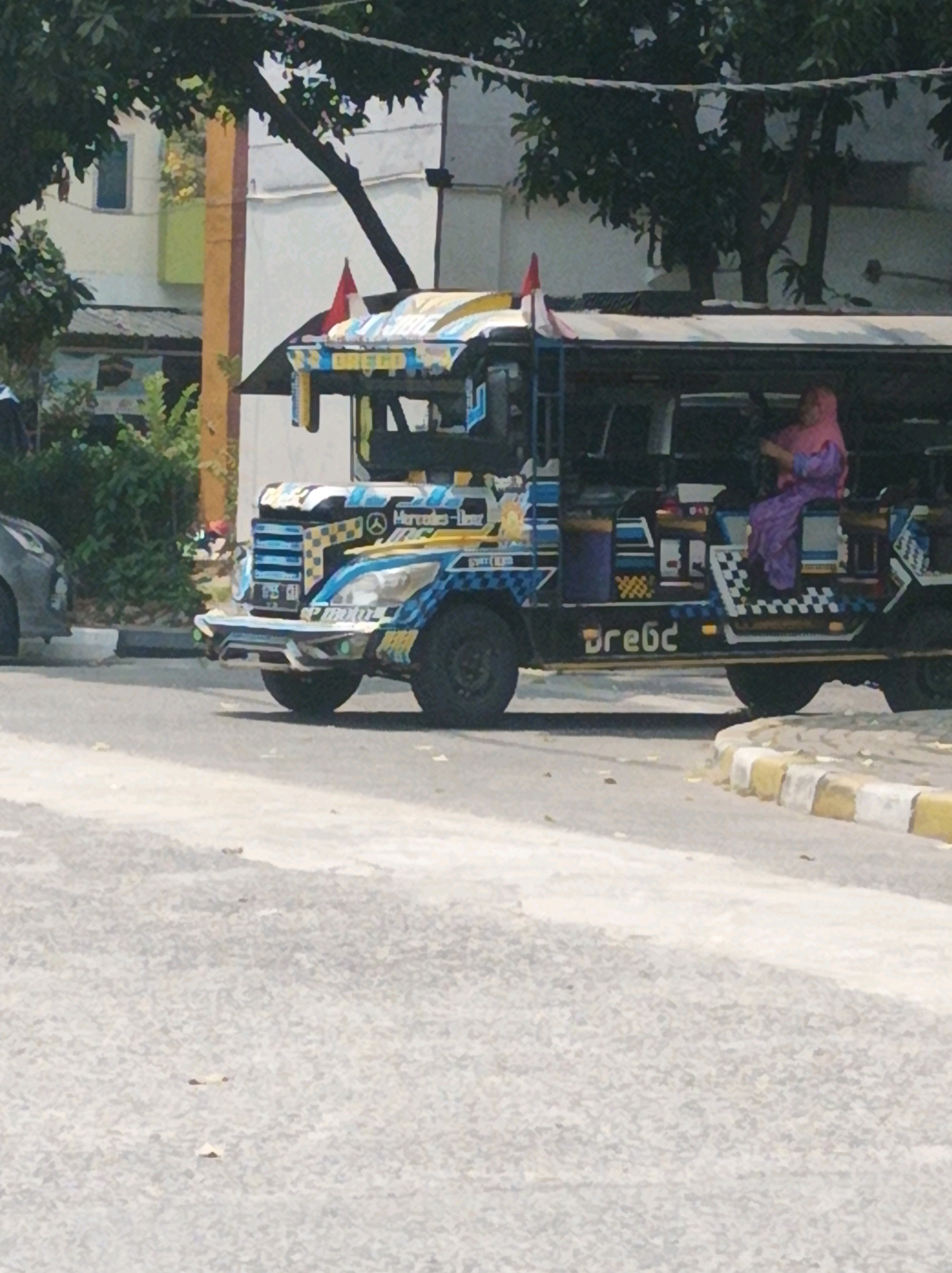In West Jakarta, Indonesia, a colorful jeepney, a public utility vehicle common in the Philippines and some parts of Indonesia, is parked or driving on a street. The jeepney is decorated with various patterns and colors, including blue, yellow, and white, with checkered accents. A person wearing a pink headscarf and a purple dress is visible inside the jeepney, seemingly driving. The environment suggests a street scene with buildings and trees in the background. The road has a concrete curb with yellow and white markings. The overall impression is of a vibrant and busy urban setting.