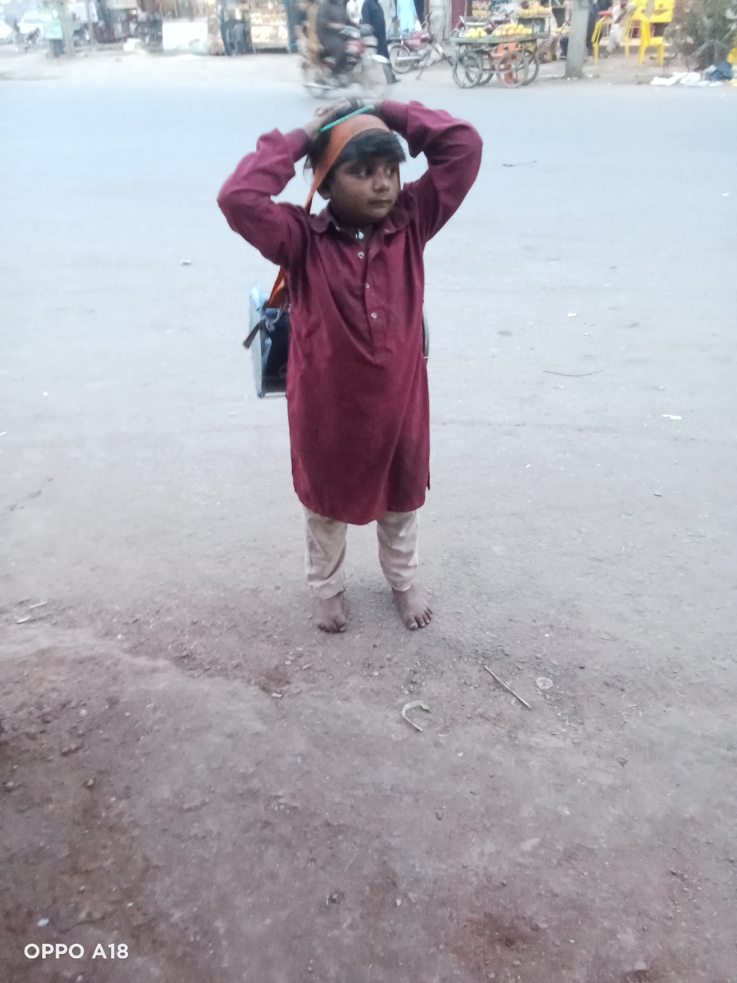 A young boy stands barefoot on a dirt and asphalt road in an outdoor setting. He appears to be around 6-10 years old, dressed in a maroon, long-sleeved kurta and light-colored pajama bottoms. A brown or orange fabric band is wrapped around his head, and his hands are raised, resting on or near the back of his head, possibly adjusting it. A small, dark backpack or satchel is visible on his back. He faces slightly to his right, looking intently towards something outside the frame.
The background reveals a bustling street scene with blurred figures, motorcycles, and street vendors, including a pushcart that might be selling fruit. The ground is dusty, with small pieces of debris like sticks and a bent metal object. The lighting is diffused, suggesting either an overcast day, early morning, or late afternoon. The overall environment indicates a busy urban or semi-urban area, possibly in a developing country, characterized by modest infrastructure and street-level commerce. A watermark "OPPO A18" is present in the bottom left corner of the image.