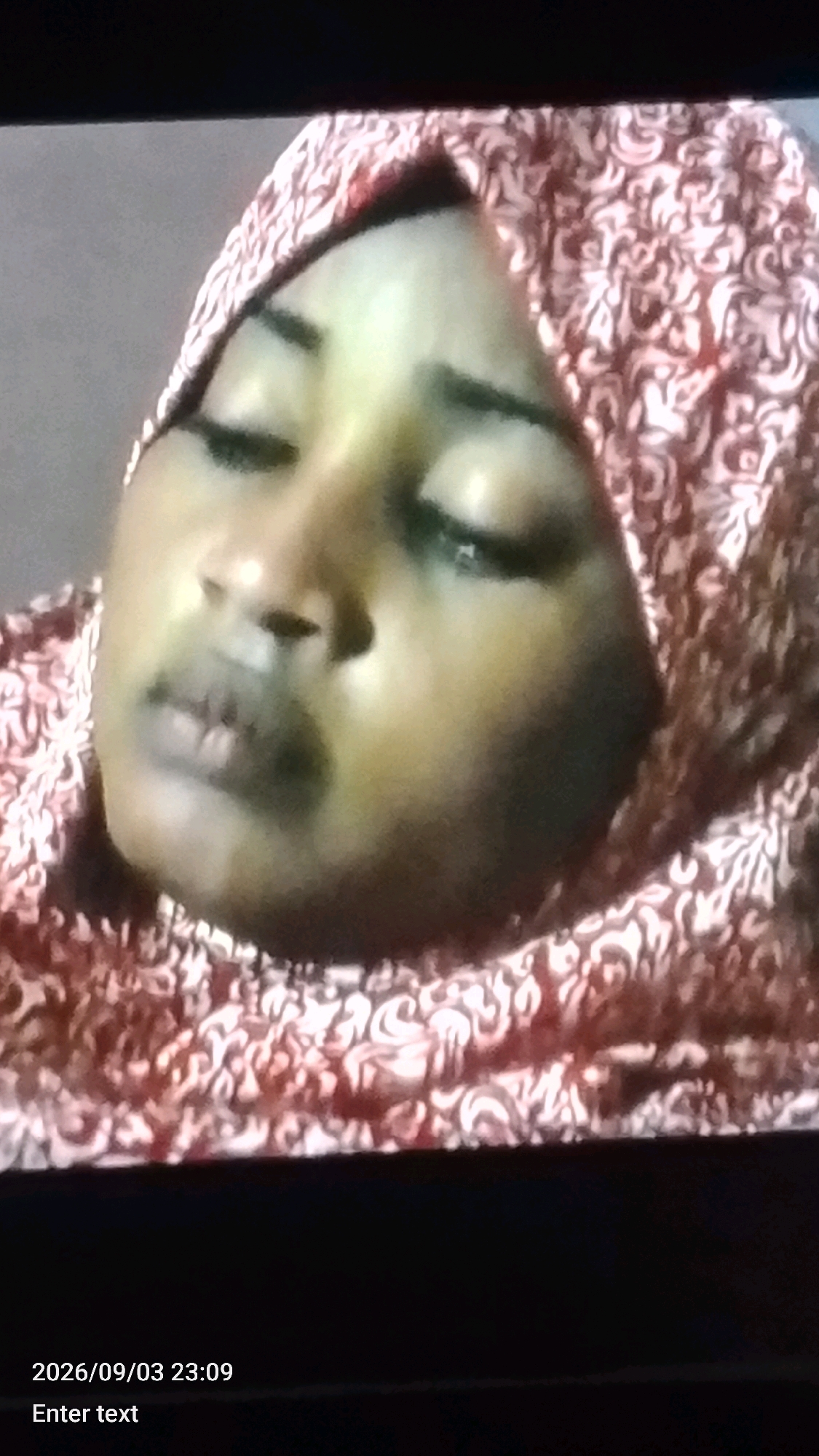 A close-up shot captures a woman wearing a patterned headscarf. The woman's eyes are closed, and her head is tilted slightly downwards. Her facial features are visible, including her nose and lips. The headscarf is red and white with an intricate pattern. The background is dark and out of focus, suggesting the image was taken in low light conditions. The bottom of the image displays a timestamp "2026/09/03 23:09" and the text "Enter text". The image does not provide any contextual information about the location in Lokoja, Nigeria.