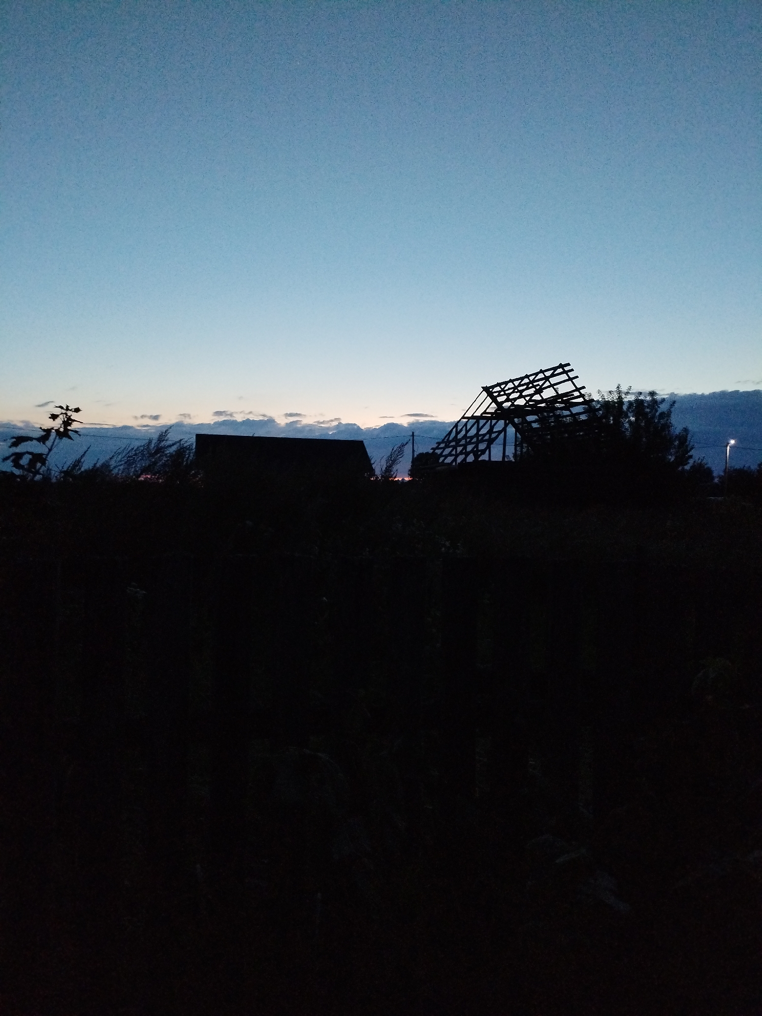 The image depicts a twilight scene in Bialynichy, Belarus. The sky is a gradient of pale blue to a darker shade of twilight. In the foreground, a dark, silhouetted landscape is visible, featuring dense vegetation and the outlines of what appear to be farm buildings. One prominent structure is a building with a partially constructed or ruined roof, exposing its wooden frame against the sky. To the right, a street lamp emits a faint yellow light. The overall impression is one of a rural setting transitioning from day to night, with signs of human habitation and potential construction or decay.