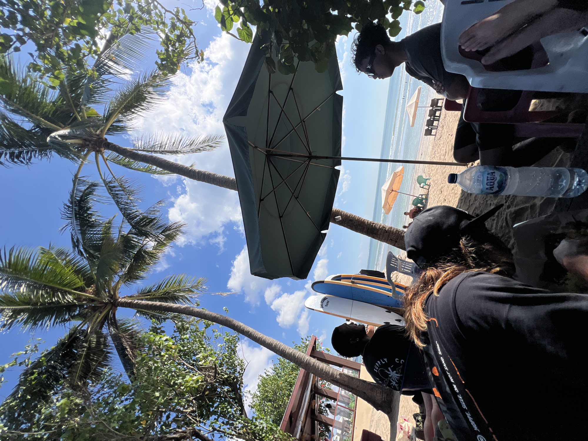 This image depicts a sunny day at Kuta Beach in Bali, Indonesia. The foreground is dominated by beachgoers seated on the sand under a large green umbrella, with palm trees providing shade. Several surfboards stand upright nearby, suggesting a surfing activity. In the background, the ocean stretches to the horizon under a bright blue sky with scattered clouds. The beach is lined with lounge chairs and umbrellas, indicating a popular tourist spot. The overall atmosphere is relaxed and summery.