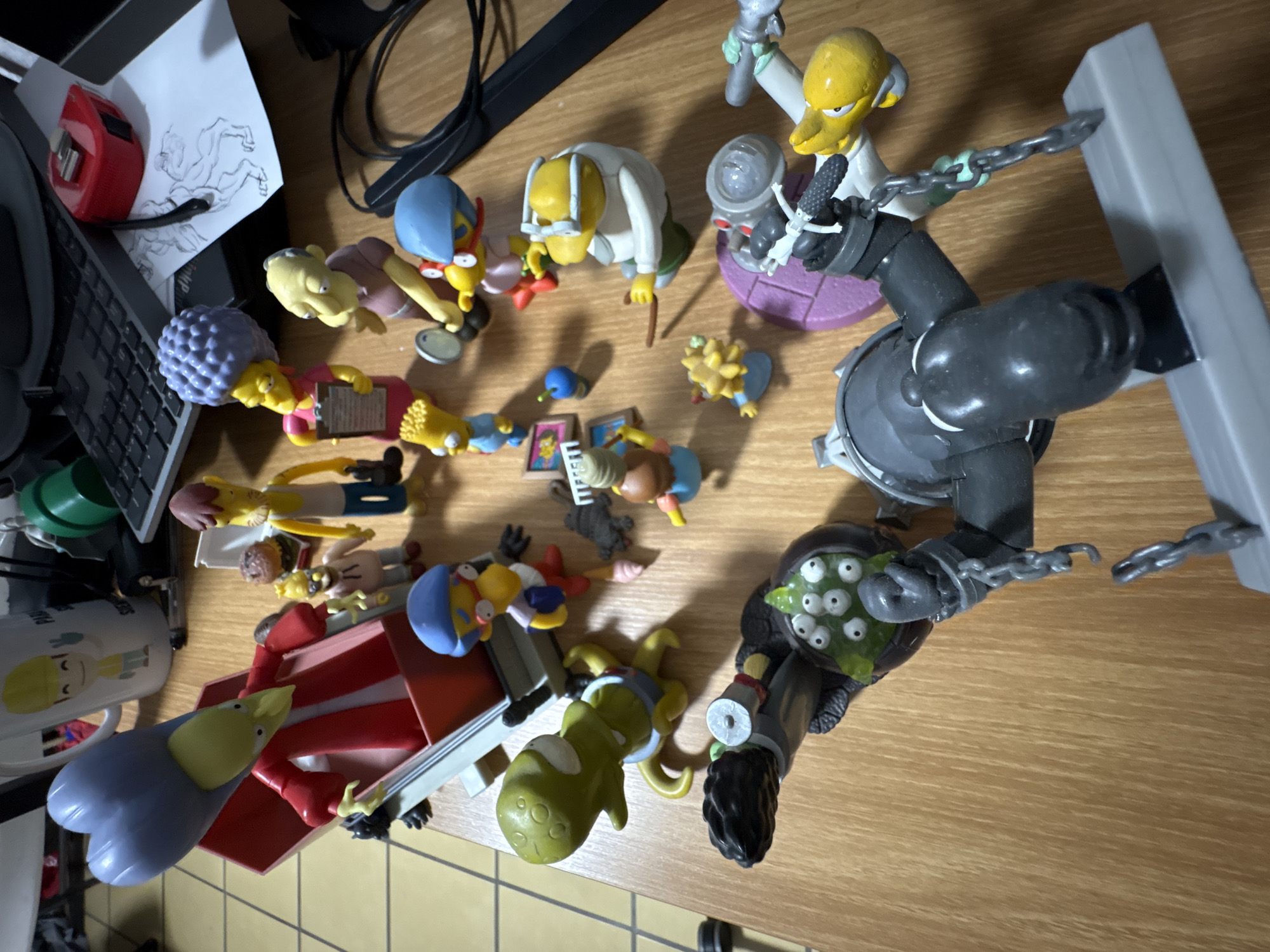 The image depicts a collection of Simpson's figurines spread across a wooden desk. The characters present include: Mr. Burns with Smithers, Homer Simpson, Marge Simpson, Bart Simpson, Kang, Kodos, and the Flying Hellfish. Various objects such as a keyboard, a drawing, a mug with the Simpsons on it, and loose cables are also present. It is not possible to determine the specific location in Cuernavaca, Mexico from the image.
