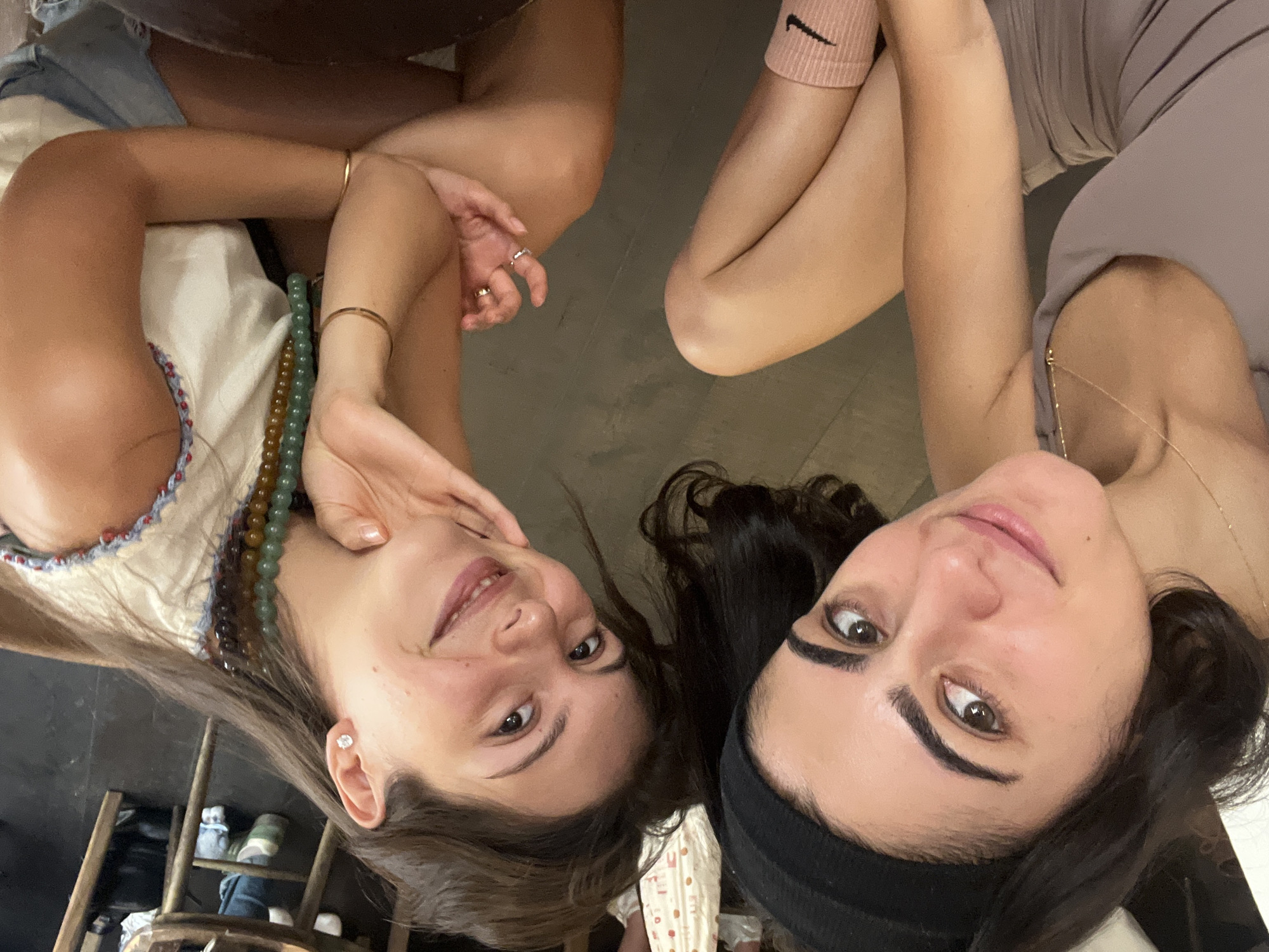 Two young women are shown in a close-up, upper-body selfie. The woman on the left has dark hair with a black headband and is wearing a taupe tank top and pink socks with a black Nike logo. The woman on the right has brown hair and is wearing a white top with blue and red trim and denim shorts. She has multiple necklaces made of beads and wears a diamond stud earring. Both women are smiling and appear to be in a casual, indoor setting. The background includes wooden furniture, suggesting a cafe or restaurant. There is no indication in the image that this location is in Paris, France.