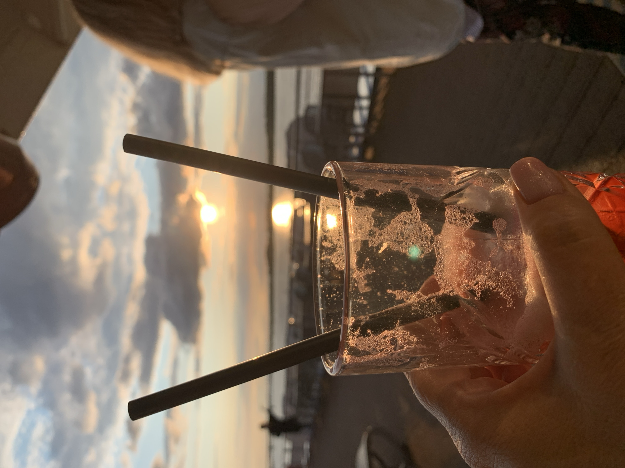 The image shows a close-up of a hand holding a glass with two straws in it, in front of a sunset over a body of water. The glass contains a pinkish liquid with some foam and condensation. The background features a sky with clouds illuminated by the setting sun, and a distant silhouette of a person standing on a railing. A blurred figure of another person is visible on the right side of the frame. The scene appears to be on a deck or walkway, with wooden planks visible in the foreground and background. There is no discernible information in the image to suggest a specific location in the city of Perm, Russia.