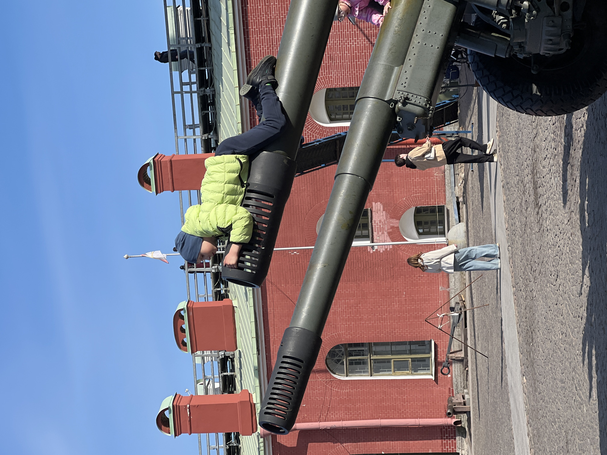 A young boy in a lime-green jacket and dark blue pants and hat is perched on a cannon barrel in the Peter and Paul Fortress in Saint Petersburg, Russia. His legs are extended upward, with feet in black shoes resting near the top of the barrel. The cannon is positioned against a red brick building featuring arched windows with multiple panes. Chimneys are visible on the building's roof. Other people are walking nearby, including a woman in a beige coat and dark pants, and another woman in a white jacket and light blue jeans. A metal structure resembling a camera stand is also present. The sky is a clear blue.