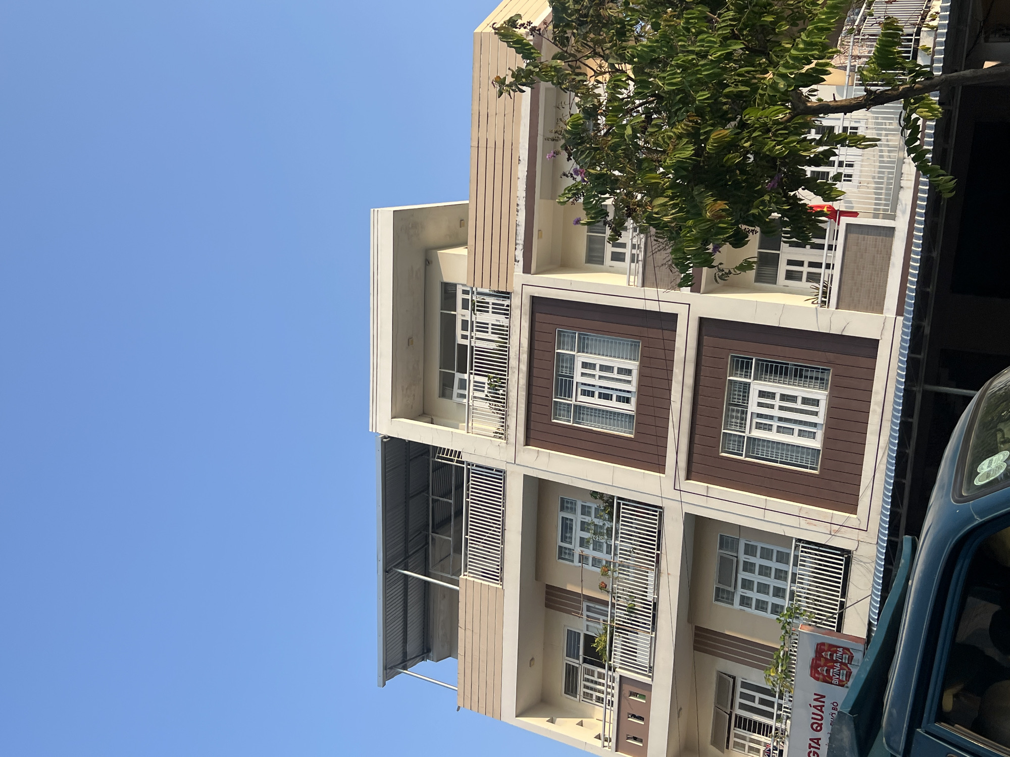 The image displays a multi-story building set against a clear, bright blue sky, indicating daytime with strong sunlight. The building features a modern design with light cream-colored walls, brown decorative panels around windows, and multiple balconies. Each level includes windows with white frames and grilles, and some balconies are fitted with horizontal slatted screens for privacy or shade.
On the upper right side, a large, leafy tree partially obscures the building, its green foliage showing hints of small purple flowers. Part of a blue vehicle, possibly a small truck or van, is visible at the bottom right of the frame, suggesting a street or parking area.
Notable location cues confirm the setting in Vietnam, aligning with the provided context of Nam Natural Heritage Area Trang, Vietnam. A small Vietnamese flag (red with a yellow star) can be seen hanging from one of the balconies. Additionally, a red and white sign, partially visible near the bottom right next to the vehicle, displays Vietnamese text including "BIA SÀI GÒN" (Saigon Beer) and "QUÁN" (Eatery or Restaurant), with another partial word "GA" also discernible. No human activity or specific events are depicted in the static scene.