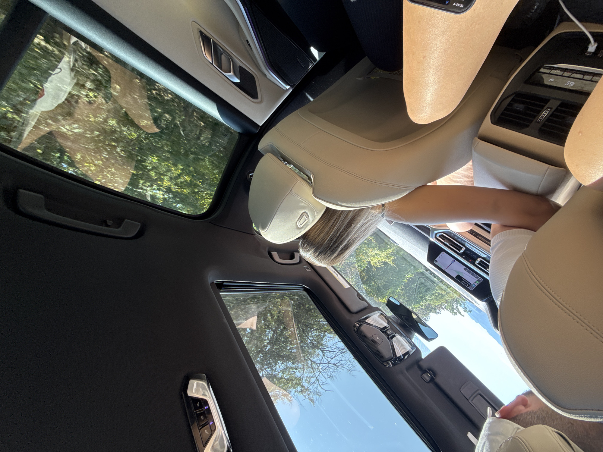The image shows the interior of a car with a panoramic sunroof open, revealing a bright, sunny day with lush green trees and a clear blue sky. The camera is positioned to capture the view from the back seat, looking forward and upward. Inside the car, the beige leather seats and the black headliner are visible. The dashboard features a large infotainment screen displaying navigation. A person's legs are visible in the foreground, suggesting they are a passenger in the car. Another person, a woman with blonde hair, is seated in front of the camera, with her back to the viewer. The scene is captured from within the car, looking out at the natural scenery passing by. There is no indication of the specific location within the city of Codlea, Romania.